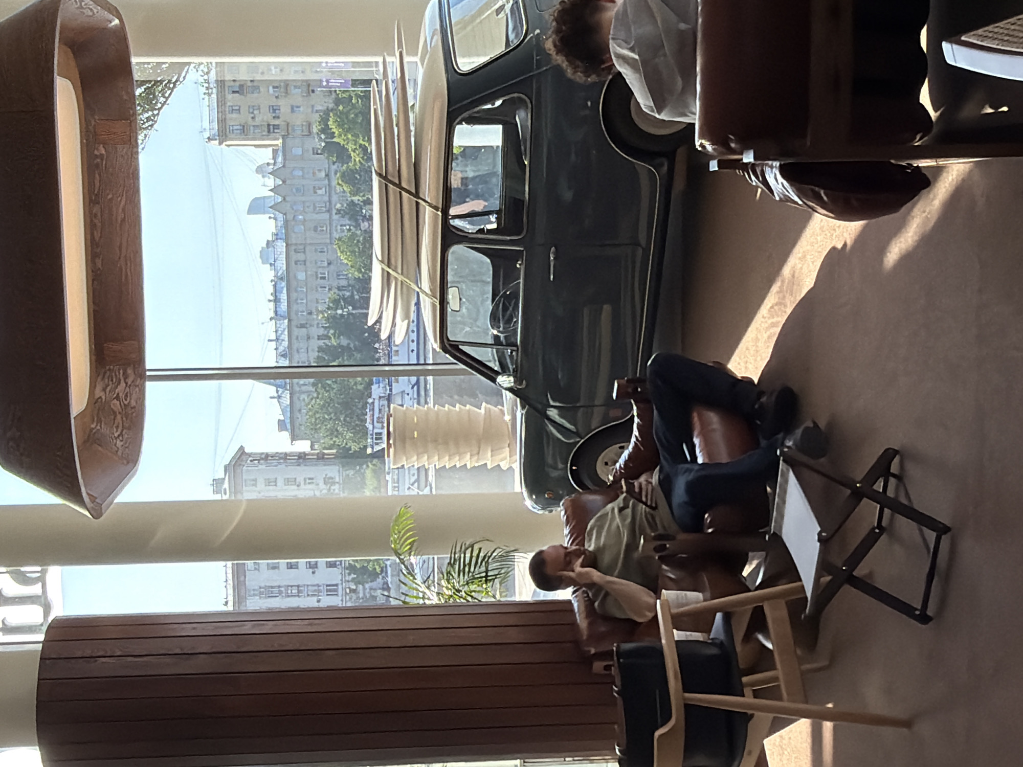 The image depicts an interior scene in what appears to be a showroom or lounge. A vintage Mini Cooper with surfboards strapped to its roof sits indoors. Two men are seated in leather chairs, one with his feet up on another chair. The setting is well-lit by sunlight streaming through a large window that reveals a cityscape of Moscow, Russia. The architectural style of the buildings visible through the window, with their distinct Soviet-era design, confirms the location. Furniture includes a wooden slat structure and modern chairs. People seem to be relaxing in a casual, yet stylish environment.