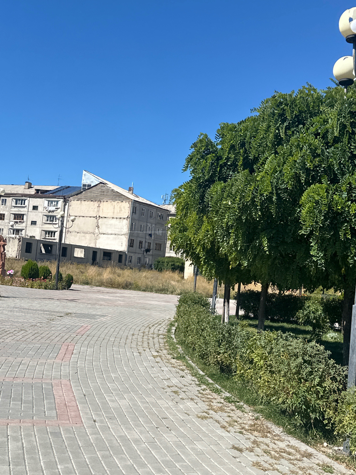 The image shows a paved walkway in a public area in Gyumri, Armenia. The walkway is made of grey and red interlocking pavers and curves to the right. On the left side of the walkway, there are some overgrown grassy areas with a few small bushes and a lamppost. In the background, there are several multi-story buildings. One building appears to be under construction or renovation, with exposed concrete and unfinished sections. Another building to its left is a lighter color with numerous windows. To the right of the walkway, there is a dense line of trees with green foliage and some smaller bushes. The sky is clear and blue.