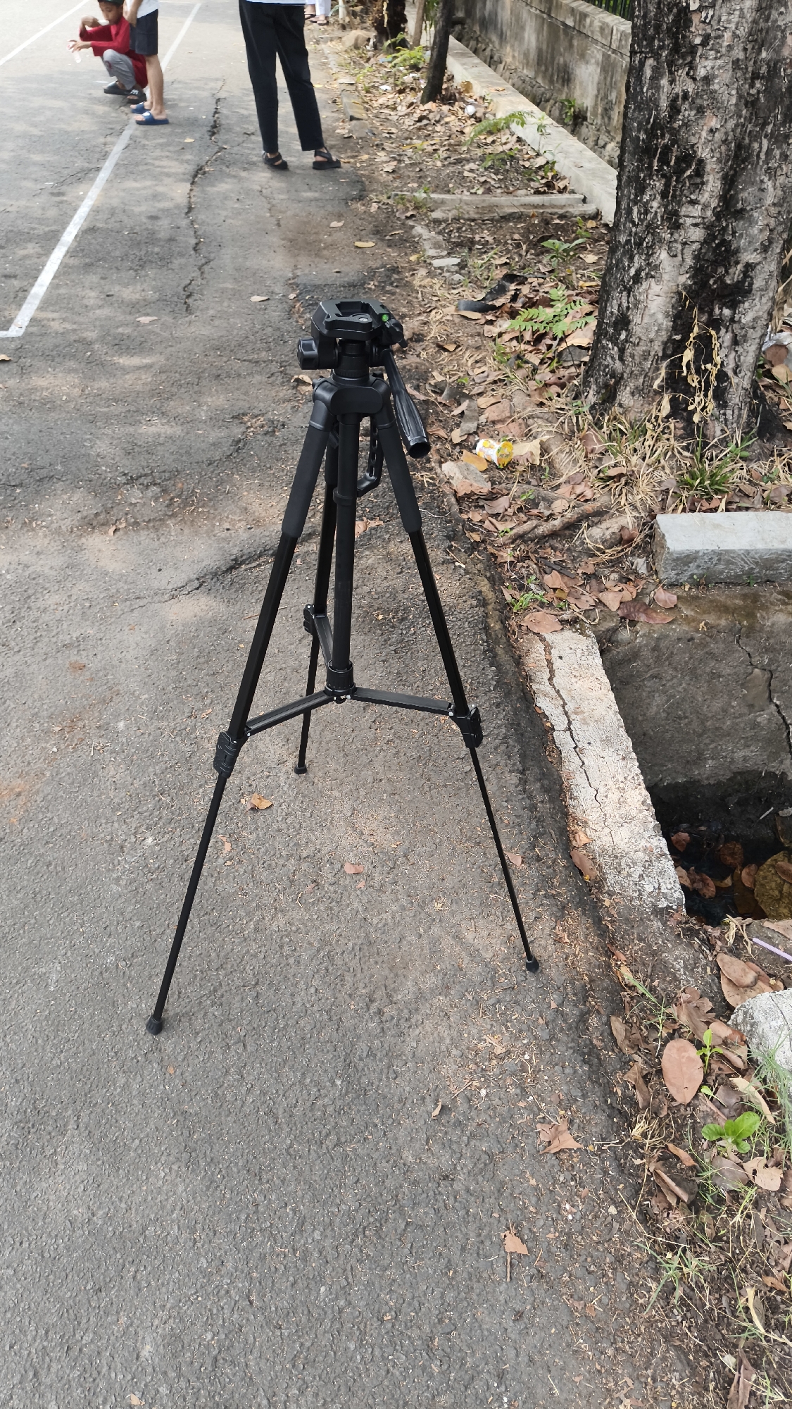 A black tripod with a camera attached stands on an asphalt surface in South Jakarta, Indonesia. The tripod is positioned in front of a large tree with rough bark and dry leaves scattered on the ground. To the left of the tripod, a white line is visible on the asphalt, suggesting it might be part of a court or sports field. In the background, several people are partially visible, including one person wearing a red shirt and another in dark pants. The environment appears to be outdoors, with some debris and vegetation present.
