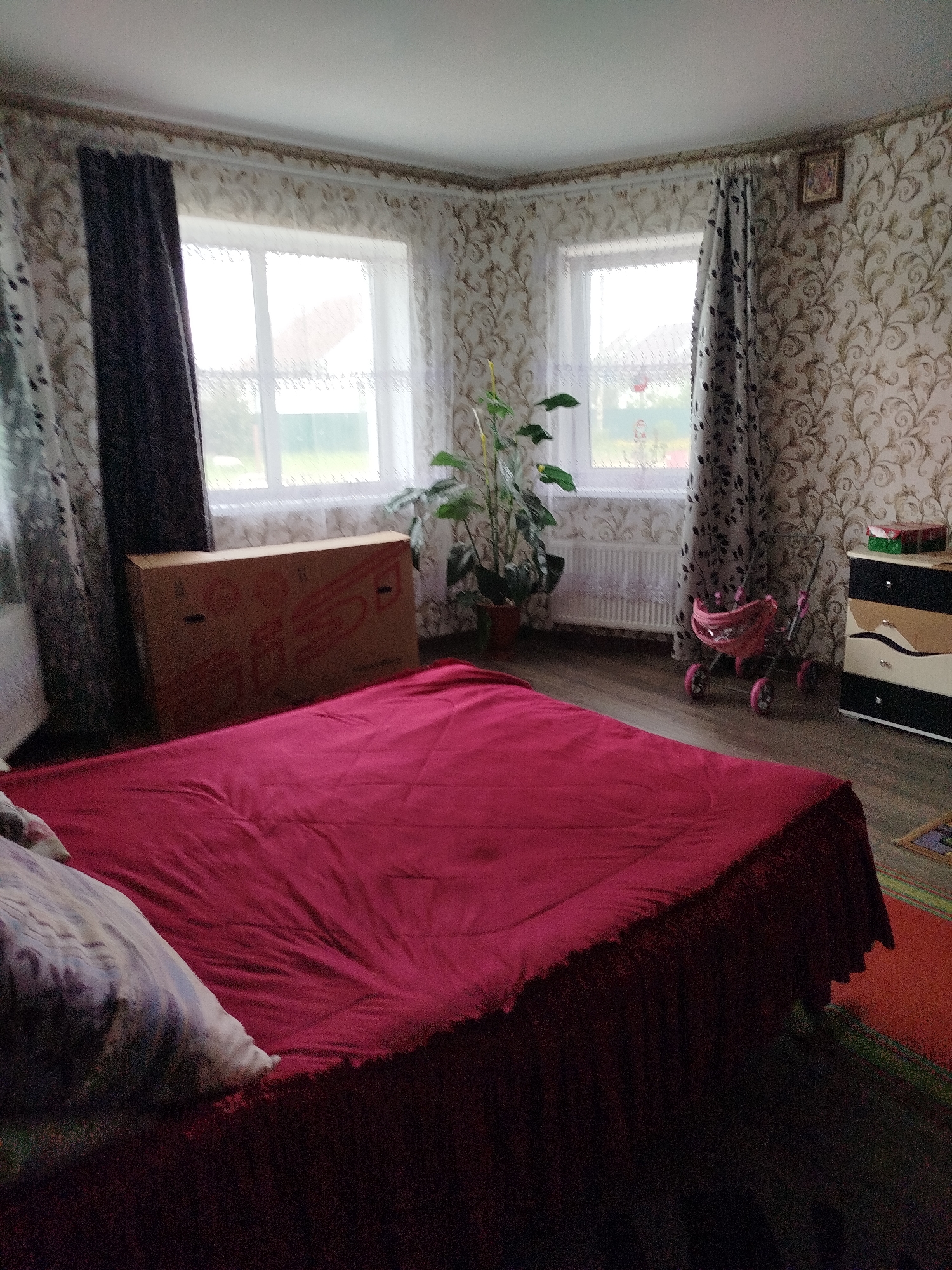 The image shows an interior scene, likely a bedroom, in a residential dwelling. The room features a large bed with a red comforter dominating the foreground. To the left, a large cardboard box is leaning against the wall. The room has two windows with patterned curtains. One window is covered by a dark curtain, while the other has a lighter, patterned curtain. A houseplant is visible near the window. In the background, a small pink doll stroller and a chest of drawers are present. The walls are decorated with a patterned wallpaper. A small framed picture hangs on the wall above the window on the right. The floor appears to be wood or laminate. The scene suggests a lived-in space, possibly with children given the presence of the doll stroller. The overall impression is one of a domestic environment.