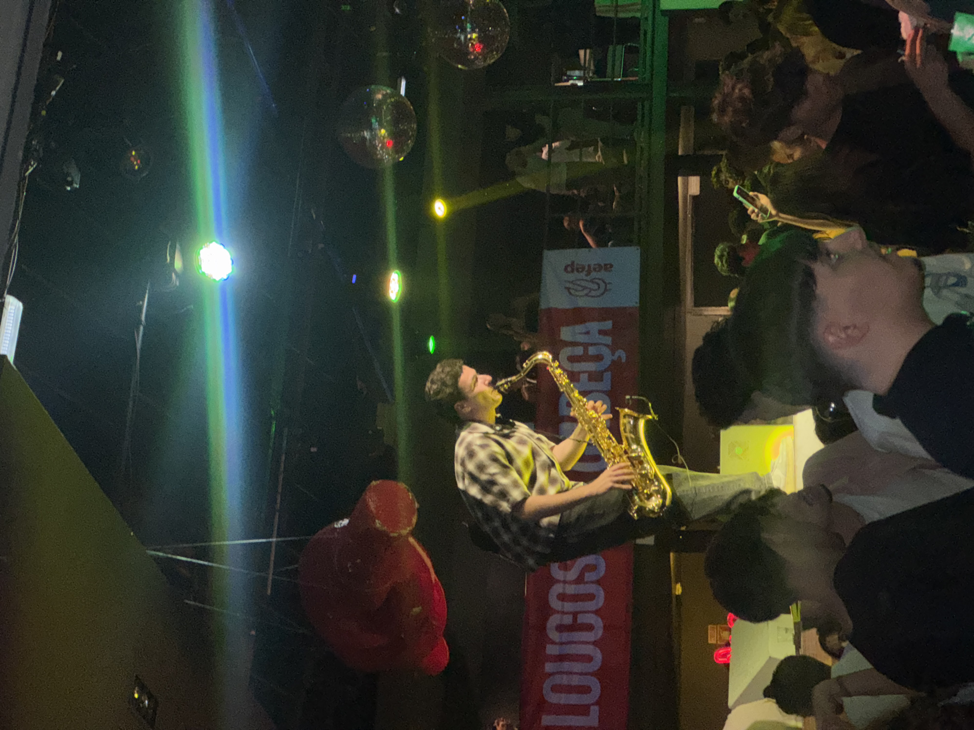 The image captures a lively scene, possibly in the city of Porto, Portugal, as suggested by the Portuguese text on the banner. A man is playing a saxophone, positioned in front of a red banner with white lettering. A large red object, possibly a decoration, is to the left. Disco balls and colorful lighting add to the ambiance. On the right, a crowd of people appears to be enjoying the performance, with some holding up phones, possibly recording or taking pictures.