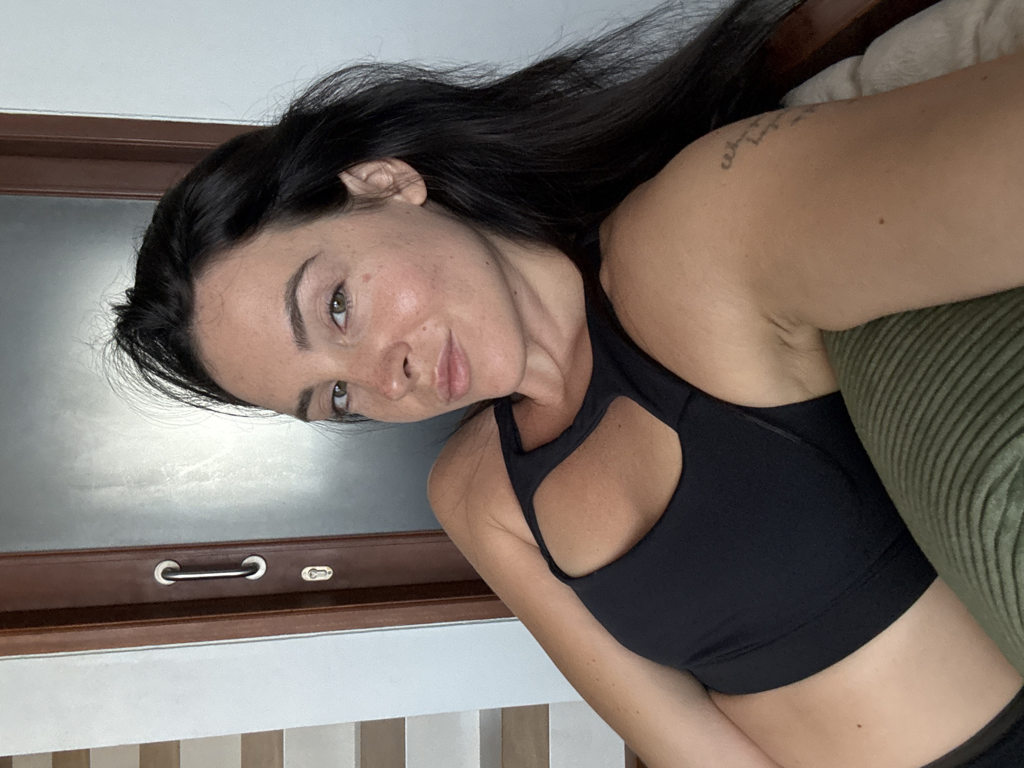 A woman with long, dark hair and light skin is captured in a selfie. She is wearing a black sports bra and appears to be indoors, possibly in a hotel room in North Kuta, Indonesia, given the presence of a wooden door with a handle and lock, and a wall that could be part of a hotel's interior design. Her gaze is directed towards the camera with a relaxed expression. The background suggests an indoor setting with architectural elements like a door frame and what looks like a vertical blind or shutter system.