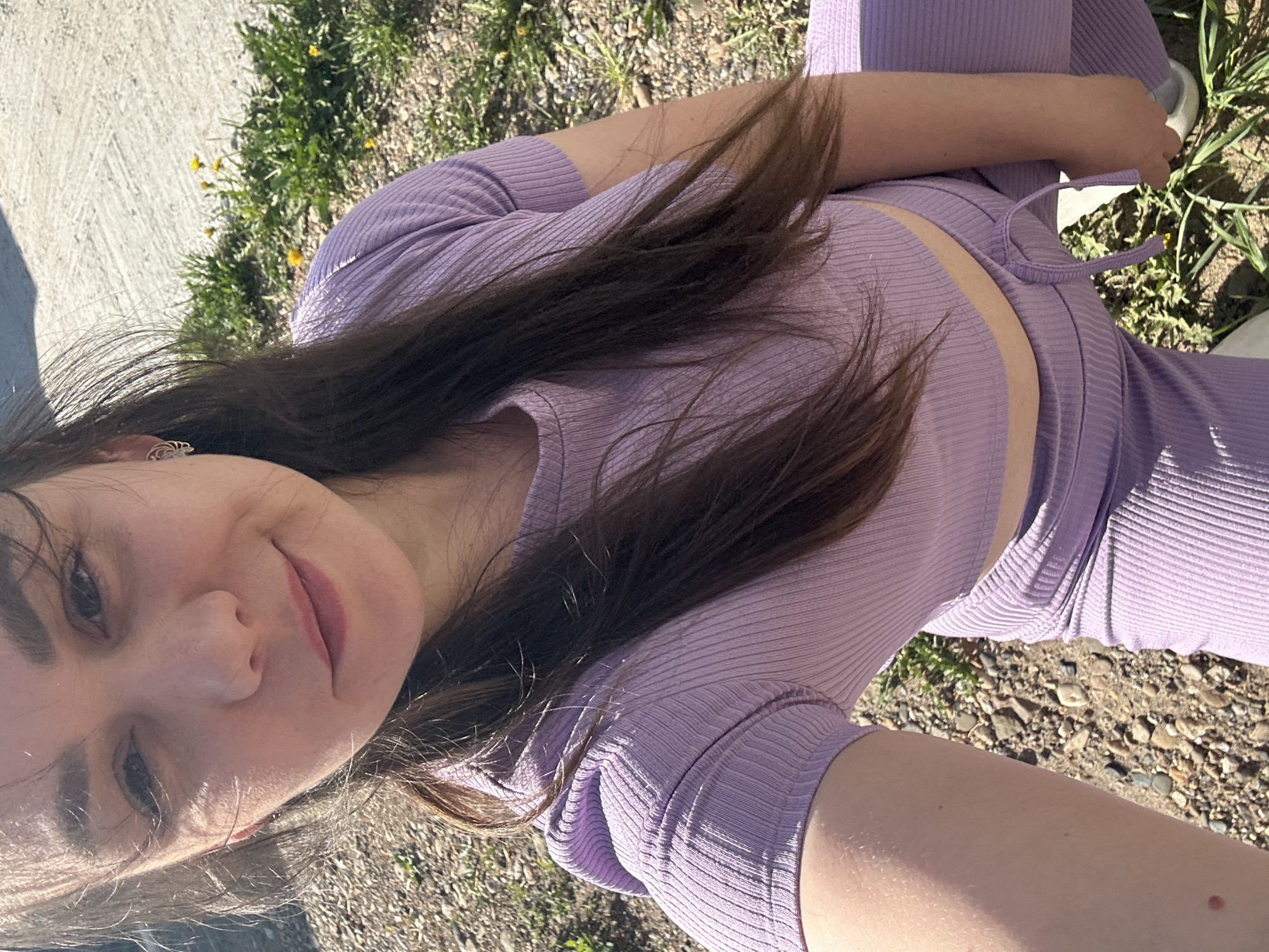 A young woman with brown hair is shown from a close, overhead perspective. She is wearing a lavender-colored ribbed two-piece outfit consisting of a short-sleeved top and pants with a drawstring waist. The background consists of patches of grass and dirt, and a concrete sidewalk is visible to the left. The setting appears to be outdoors, possibly in a park or green space. The geographic location is Chaikovsky, Russia, however, no location-specific elements are noticeable in the image.