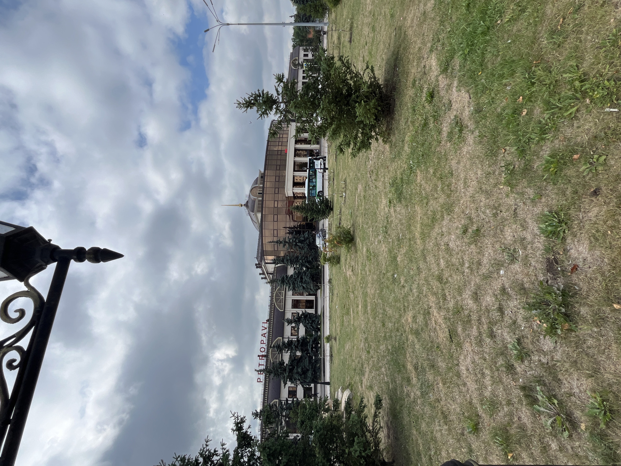 A wide shot shows a cloudy day in front of the Petropavl train station in Petropavl, Kazakhstan. The train station is a large building with a modern facade of glass and some ornate architectural elements. Red lettering spelling "PETROPAVL" is visible above the entrance. Several fir trees are planted in front of the building, and a grassy area with some scattered weeds is in the foreground. A bus and a car are parked in front of the station, and a street lamp stands to the right of the building. A decorative lamppost with a curled metal design is visible in the upper left corner of the frame, partially obscuring the sky.