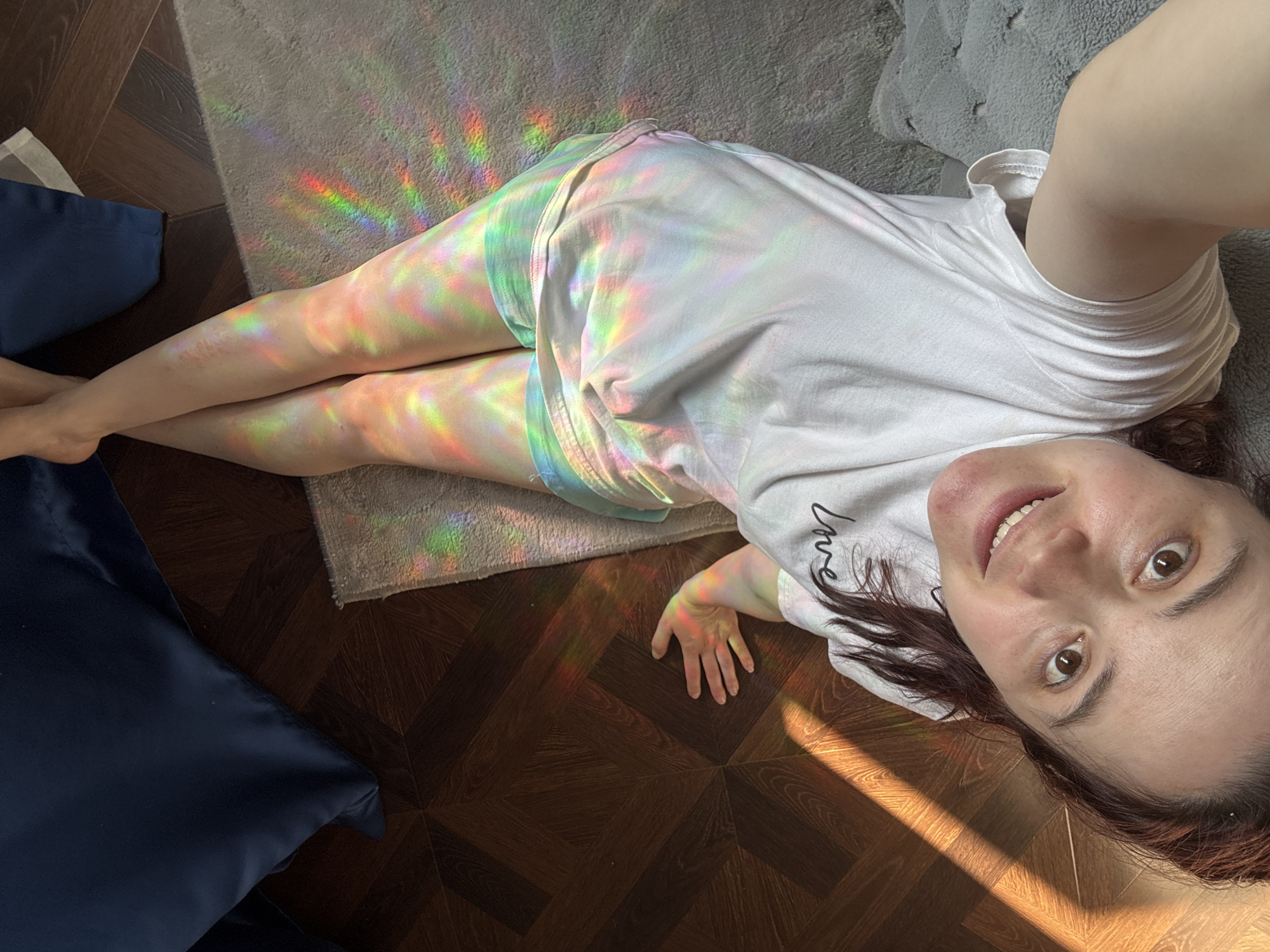 A young woman is lying on the floor taking a selfie. She is wearing a white t-shirt and shorts, and her legs are illuminated by a rainbow prism effect. The floor is a dark brown parquet wood, with a gray rug underneath part of the woman's body. There is a dark blue cushion in the lower left corner of the frame and a gray cushion in the upper right. Part of the woman's face is visible in the upper right of the frame; she is smiling. There is also natural sunlight visible on the floor next to her. There is no information to indicate this photo was taken in Sochi, Russia.