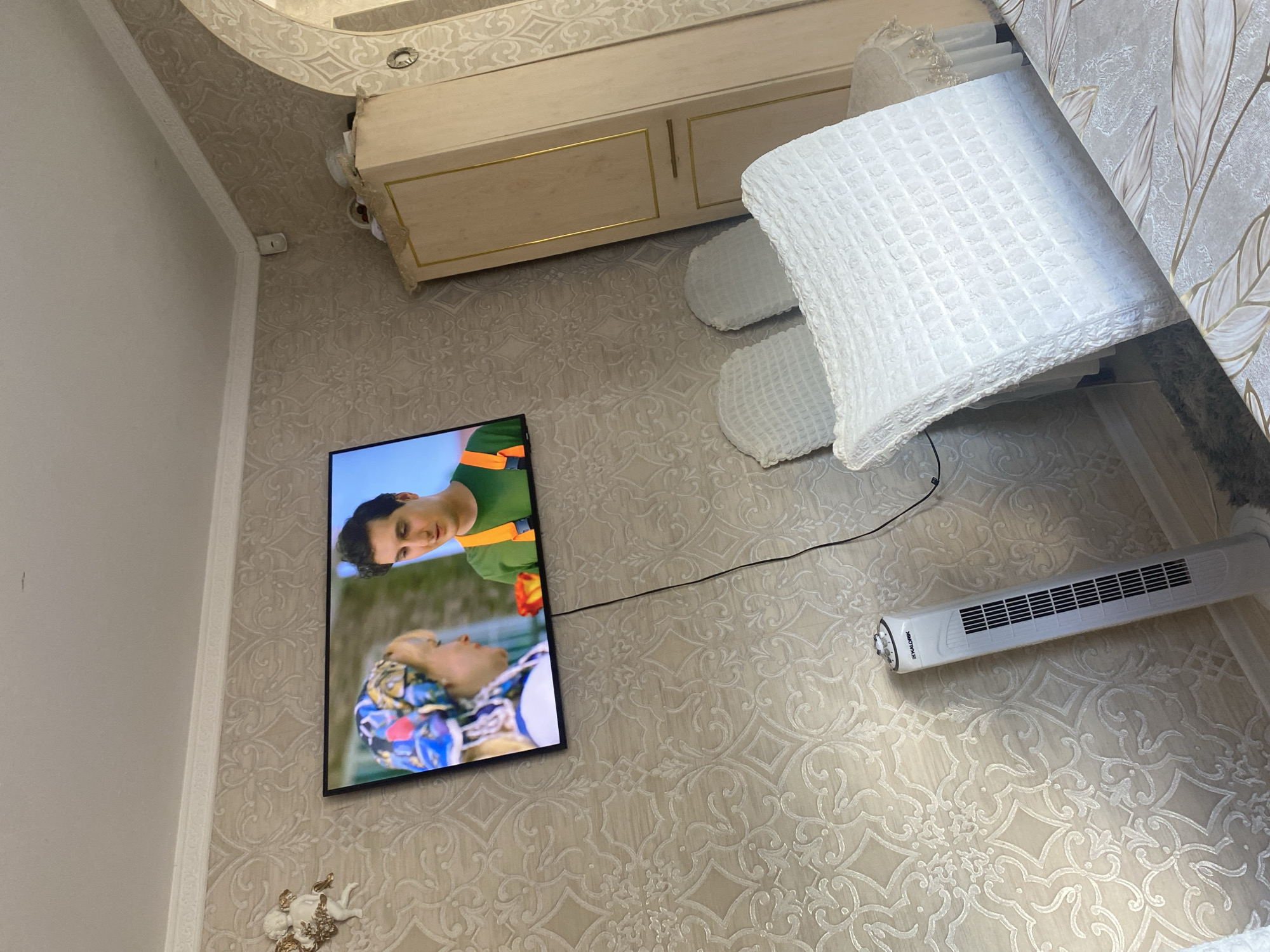 The image depicts an indoor scene, likely a living room, with a television mounted on a wall covered in textured wallpaper. On the TV screen, two individuals are visible: a woman wearing a colorful headscarf and a man in an orange harness over a green shirt. In the foreground, a white tower fan stands vertically. Behind the fan, a tall, light-colored wardrobe with gold trim is partially visible. Two white chairs, draped with textured covers, are positioned in front of the wardrobe. The overall environment suggests a domestic setting. There is no identifiable information or visual cues within the image that would allow for its specific location within the city of Daugavpils, Latvia.