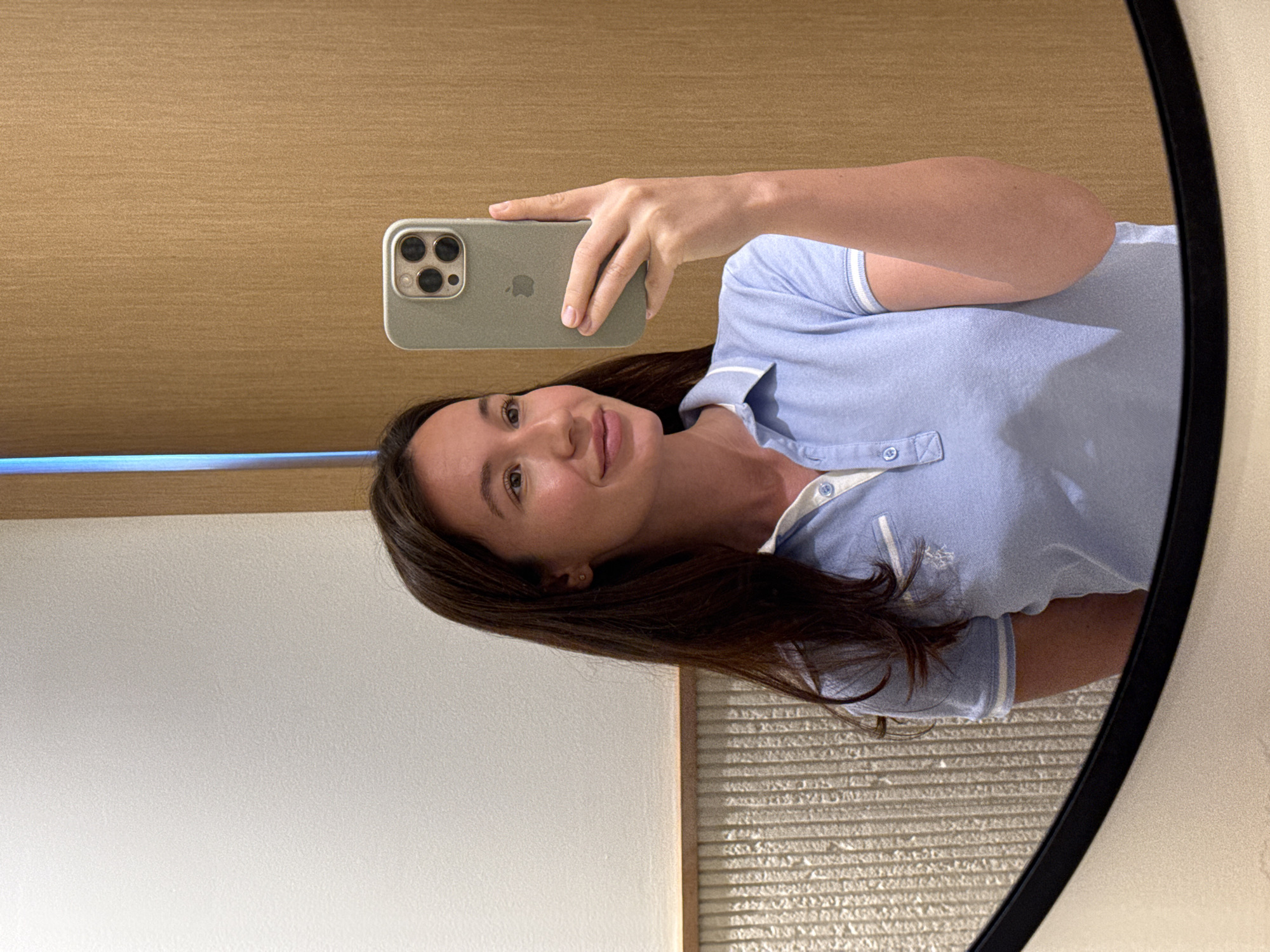 A female individual is depicted taking a mirror selfie in Tel-Aviv, Israel. She has long, dark brown hair and light skin, wearing a light blue polo shirt with a white collar and sleeve trim, which includes a small embroidered logo on the left chest. A small, gold-colored stud earring is visible in her right ear. The subject holds an olive green-cased iPhone, identifiable by its three-lens camera module, to capture her reflection. The background, visible in the mirror, shows a textured white wall on the left, horizontally segmented by a wooden trim above a vertically ridged light-colored wall. On the right, a vertically textured light brown wall is present. A vertical strip of blue light is reflected in the upper right background. The bottom edge of the mirror is framed in black, with a beige surface directly below it.