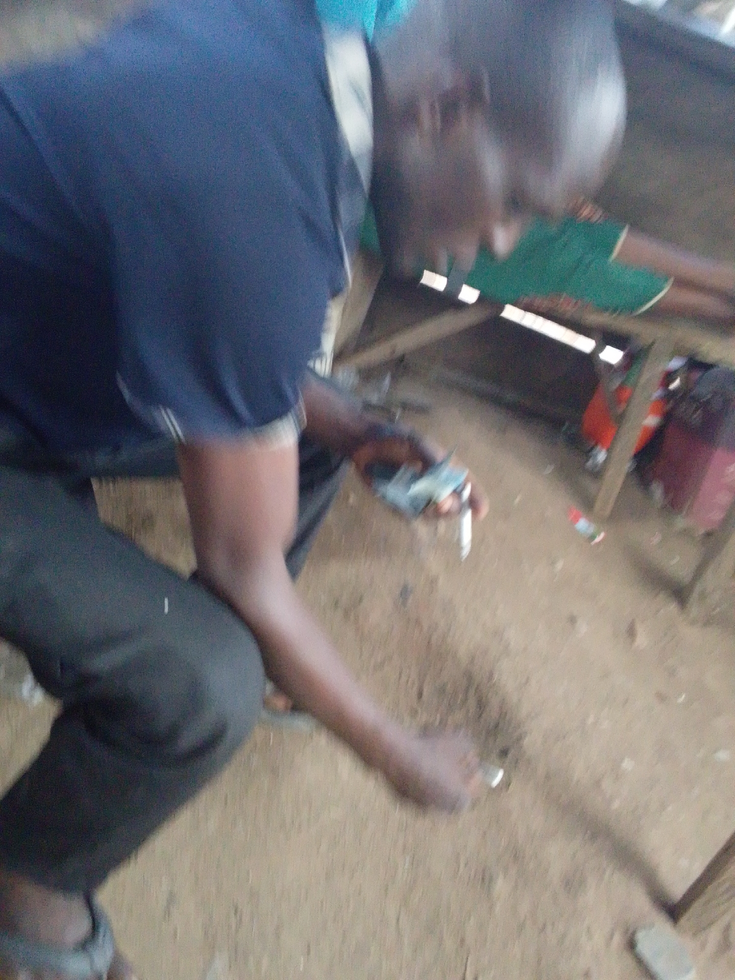 The image depicts a blurred, eye-level view of a dark-skinned man in an informal, possibly outdoor, setting. The man is wearing a dark blue short-sleeved shirt with light-colored stripes on the sleeves, and dark trousers or shorts, paired with open-toed sandals. He appears to be crouching or sitting, leaning forward. His head is partially obscured and blurred, but he seems to have a bald or closely shaven head.
In his left hand, the man is holding a small stack of what appears to be currency notes, alongside a slender, cylindrical object with a metallic or white tip, possibly a cigarette. His right hand is extended downwards, close to the sandy, light brown ground, and he might be holding a small, reflective object or interacting with something on the ground. The motion blur suggests an active moment.
The background features a simple wooden structure, likely a table or bench, with a piece of green fabric draped over it. Further back, indistinct shapes and colors, including red, orange, and maroon, are visible, possibly indicating other objects, people, or parts of a structure. A small red-capped bottle or container lies on the ground near the wooden structure. The setting, with its sandy ground and basic wooden furniture, suggests a casual, possibly market or street-side environment in Lokoja, Nigeria. The lighting indicates it is daytime. No discernible text is visible in the image.