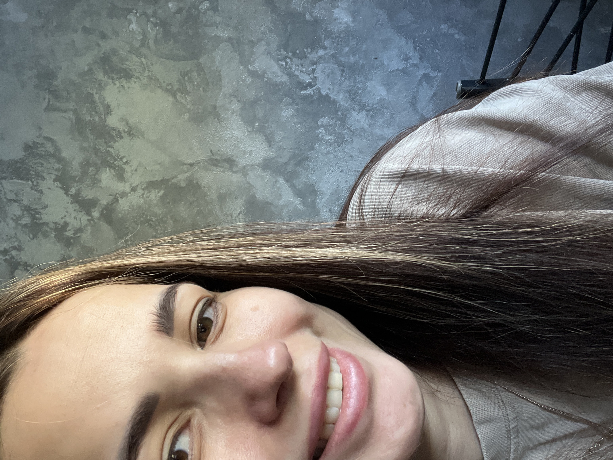 A close-up, angled shot of a smiling woman. She has brown hair with blonde highlights, brown eyes, and fair skin. Her teeth are visible. She wears a light-colored top. The background is a mottled gray wall with an unidentifiable dark object in the upper right corner. The location is Unknown, Unknown.