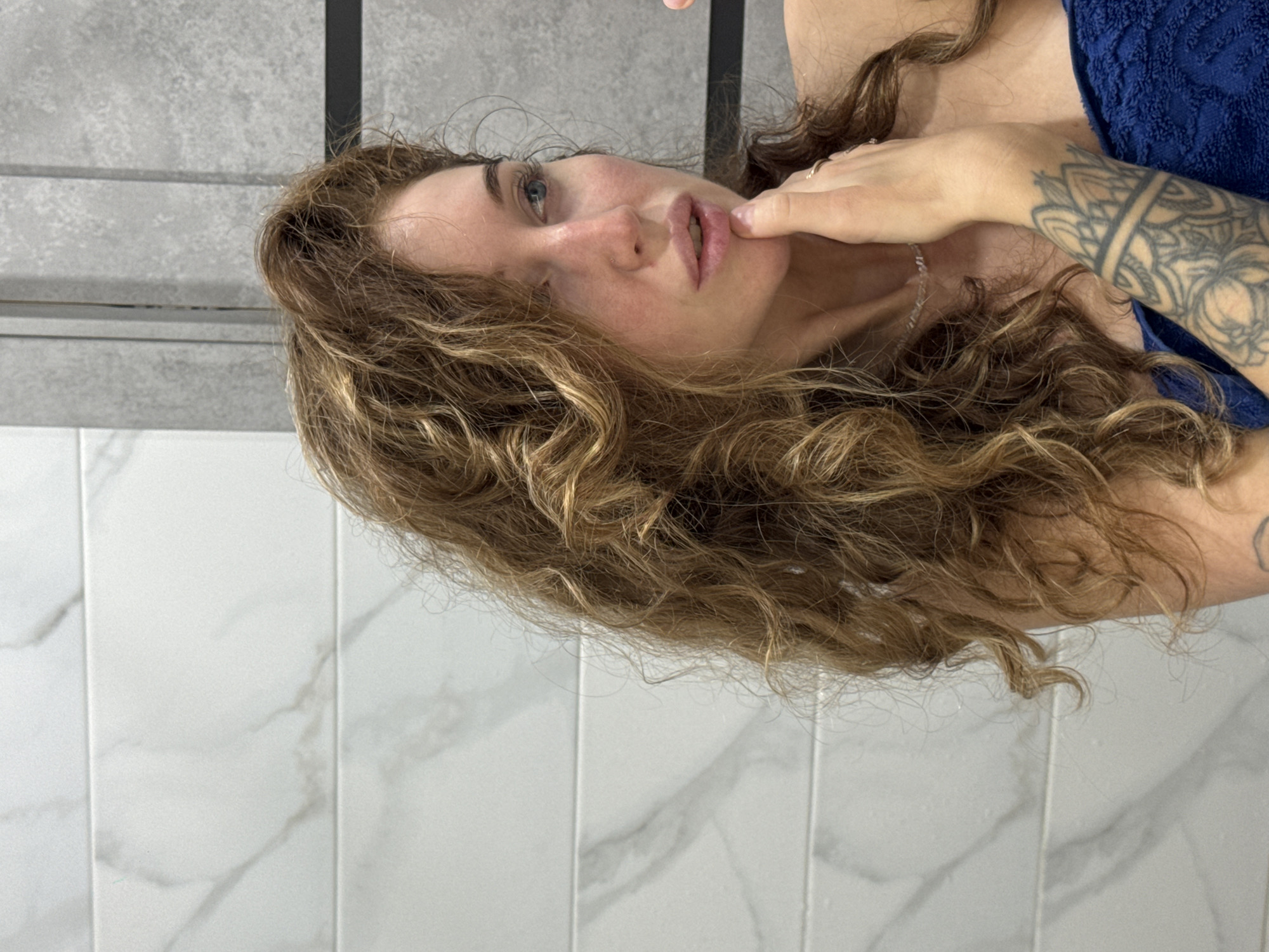 A female subject is captured in a medium close-up, gazing contemplatively to her right, likely at her reflection in a mirror. She has long, voluminous, light brown to auburn curly hair, which appears slightly damp. Her face is natural with minimal visible makeup, featuring blue eyes, a subtle nose piercing, and slightly parted lips. Her right hand is raised, with her index finger resting gently against her lower lip or chin. Rings are visible on her right hand fingers, and she wears a delicate necklace. A prominent black-ink mandala-style tattoo covers her left forearm. She is draped in a textured, royal blue towel or wrap around her upper body, covering her chest and shoulders.
The background consists of modern tiled walls. To the left, white rectangular tiles with subtle gray marble veining are arranged horizontally. To the right, gray, textured rectangular tiles are arranged vertically, separated from the white tiles by a darker vertical line, possibly an architectural feature or mirror frame. Another thin, dark horizontal line bisects the gray tiles. The specific location in the city of Chaikovsky, Russia, cannot be determined from the visual information present in the image, but the setting appears to be an indoor space, likely a contemporary bathroom or dressing area.