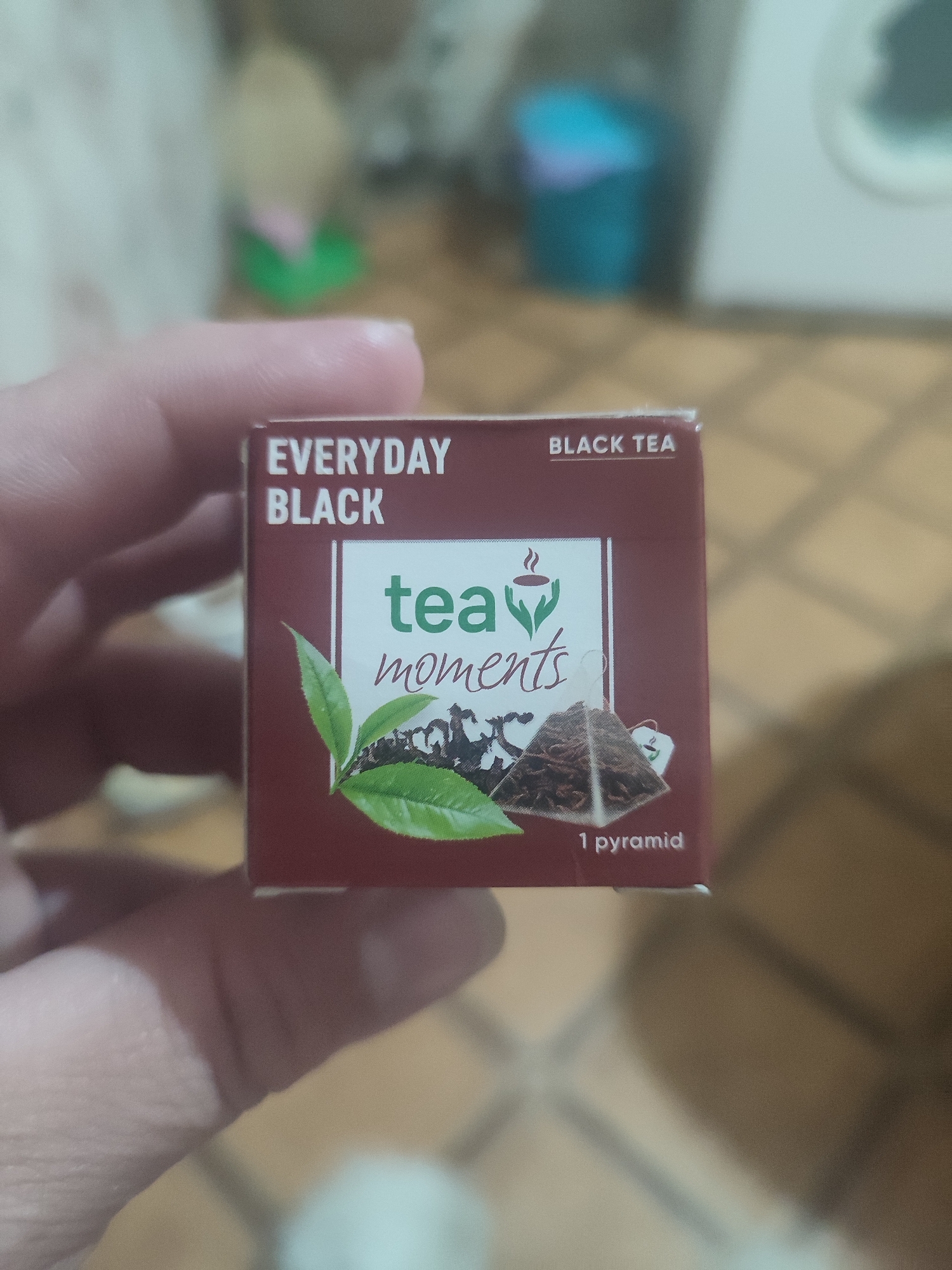 The image shows a close-up of a person's thumb and index finger holding a small box of "Everyday Black Tea". The box is dark red with white text. The text on the front of the box says "EVERYDAY BLACK" and "tea moments" with an illustration of tea leaves and a pyramid-shaped tea bag. The words "BLACK TEA" are visible on the top right corner of the box. The background is blurred and appears to be an indoor setting, possibly a kitchen or living room, with a tiled floor and some out-of-focus objects. There is no indication of the location being in Chernivtsi, Ukraine.