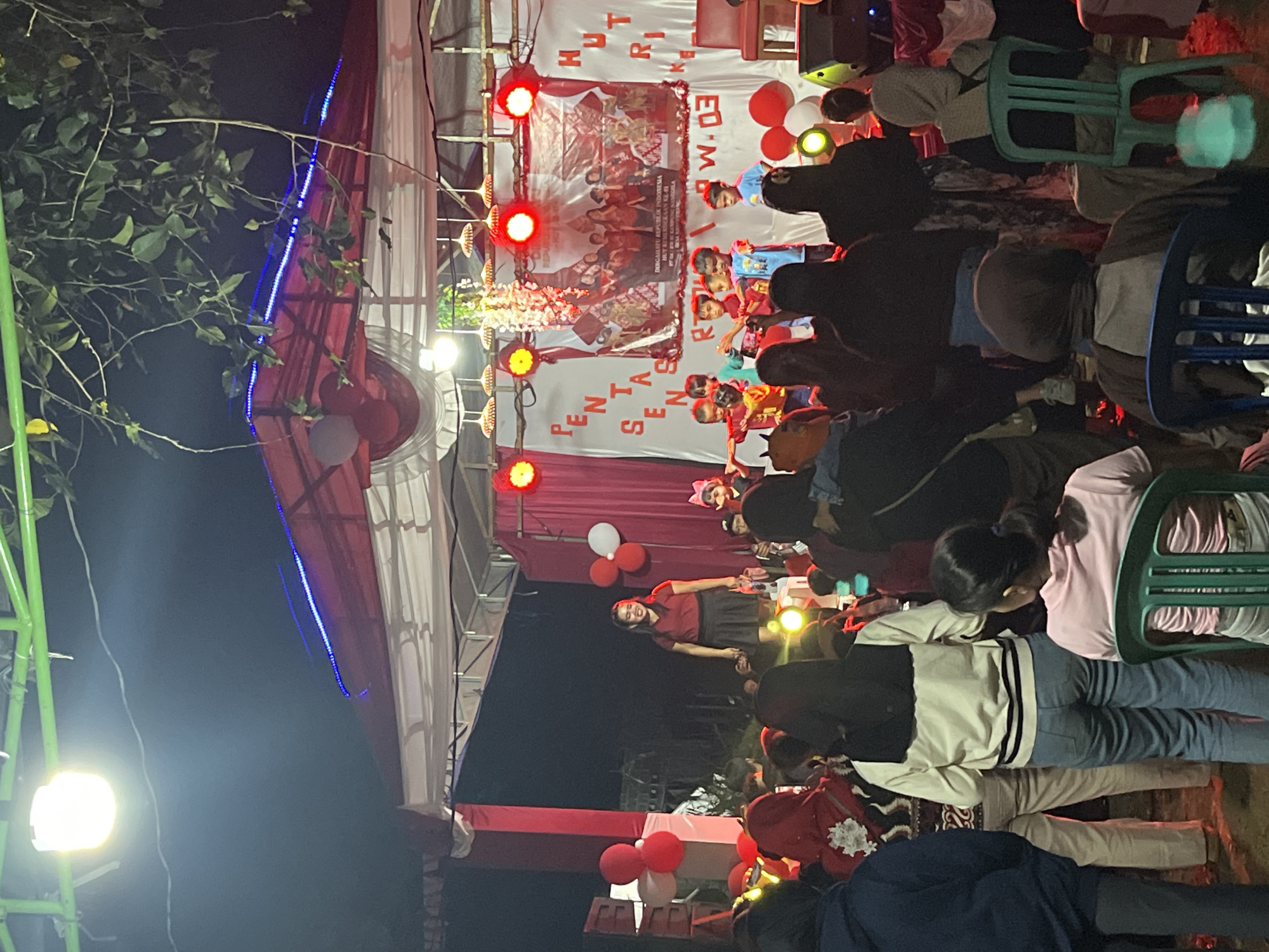 The image depicts an outdoor evening event in Tajurhalang, Indonesia, likely a community celebration or performance. A stage is set up with a red and white backdrop, featuring text that appears to be in Indonesian. Several children are on the stage, some in costume and others in casual attire, participating in what seems to be a performance or dance. A woman in a red top and black skirt is standing on the stage, possibly an instructor or performer. Red and white balloons are visible around the stage and in the foreground. A crowd of people, including adults and children, are gathered in front of the stage, watching the event. Some individuals in the audience are seated on chairs, while others are standing. The scene is illuminated by stage lights and decorative lighting. The environment suggests a casual, festive atmosphere, possibly in a public space or community center area. The overall scene conveys a sense of community gathering and entertainment.