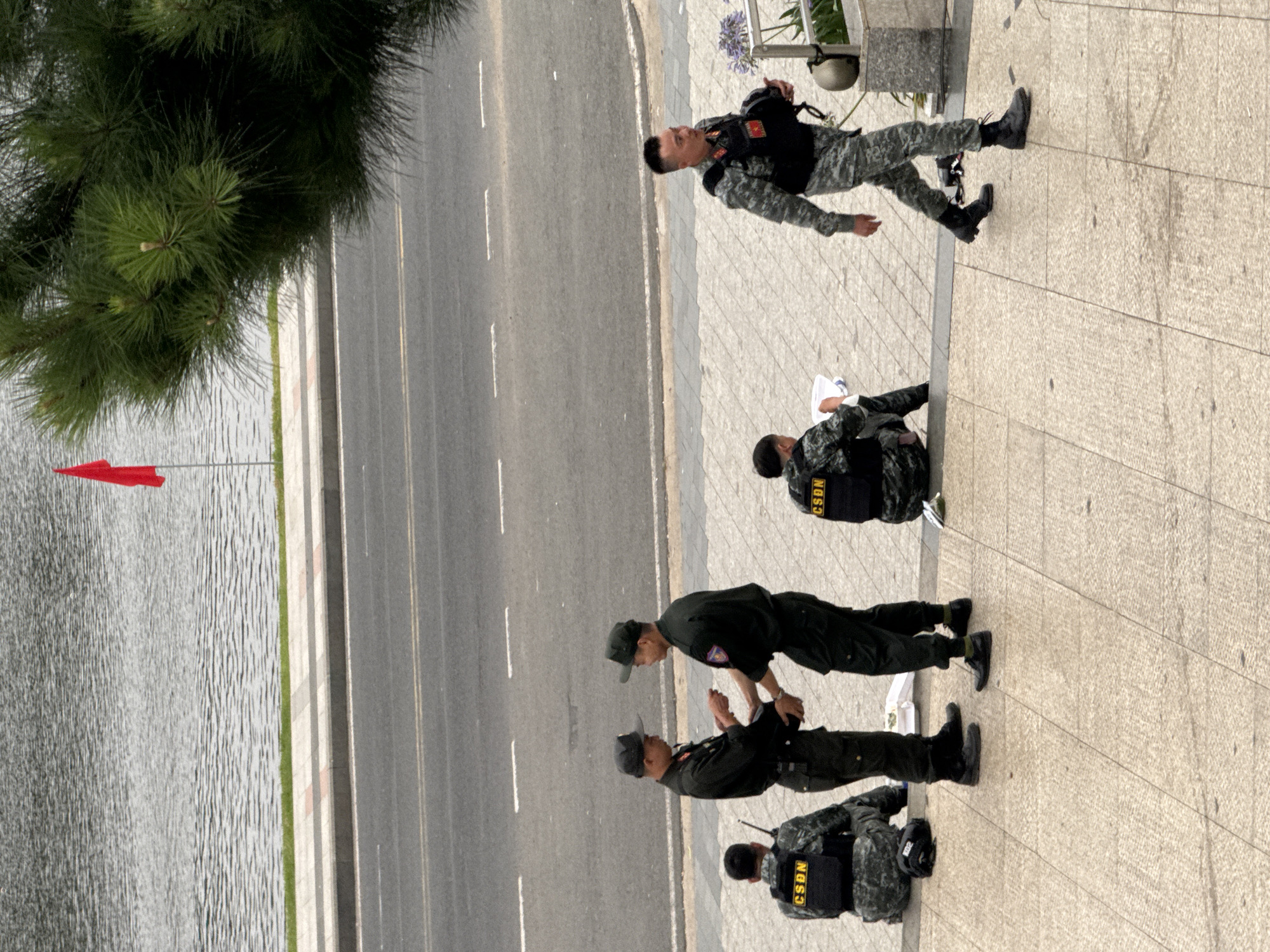 In Da Lat, Vietnam, near Xuan Huong Lake, a group of uniformed personnel are present on a paved area adjacent to a road. Four individuals are visible wearing dark uniforms and caps, with two of them interacting. One of these two is handing an object to the other. Two other individuals are seated on the ground, one facing away and wearing a vest labeled "CSDN". Another person in a camouflage uniform with a tactical vest is walking away from the group. A red flag is visible on a pole in the background near the lake. The environment suggests an outdoor public space with a body of water and a road.