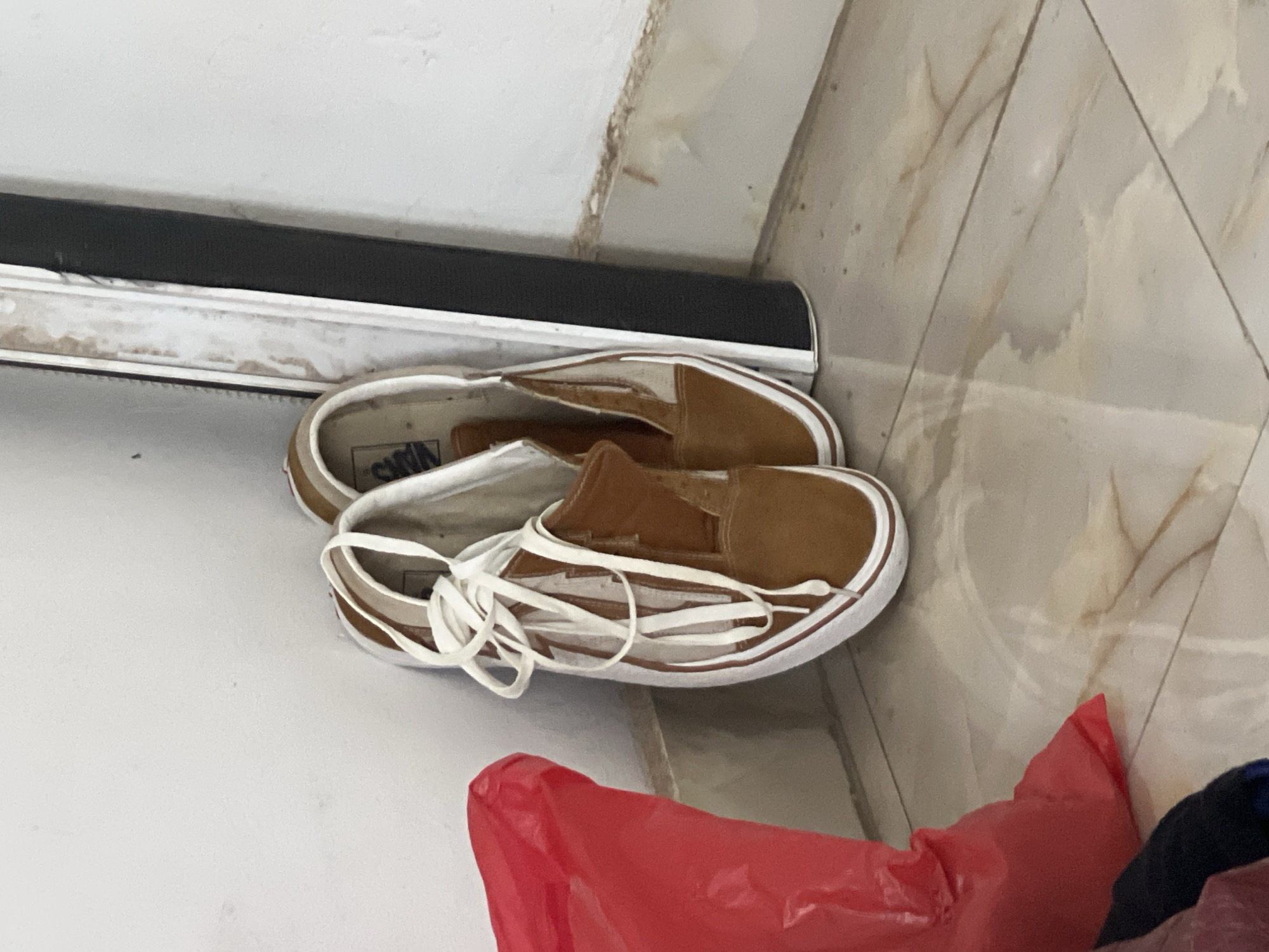 The image shows a pair of brown Vans slip-on shoes with white laces lying on a white surface near a baseboard and tile flooring. A red plastic bag and some dark blue fabric are partially visible in the lower right corner of the frame. The location cannot be confirmed as Abuja, Nigeria, without additional information.