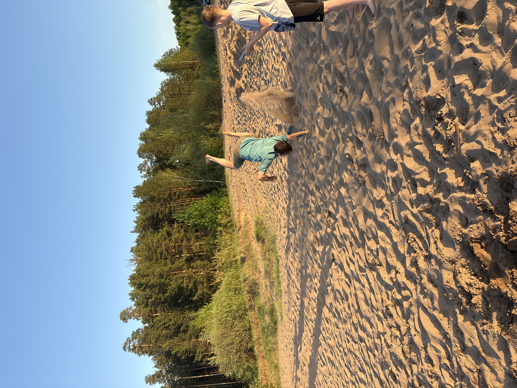 Here is a description of the image:
Location: A sandy beach in Chaikovsky, Russia, based on source metadata.
Scene: Two young males are on a sandy beach area adjacent to a line of trees and a blue sky. The sand shows footprints and other patterns.
People: One person is lying face down on the sand with arms outstretched and legs lifted, possibly in the middle of an action or fall. Another person stands near the top of the image, appearing to hold an object.
Environment: The beach is bordered by a forest or wooded area, and the sky is clear and blue. The overall lighting suggests it is daytime.