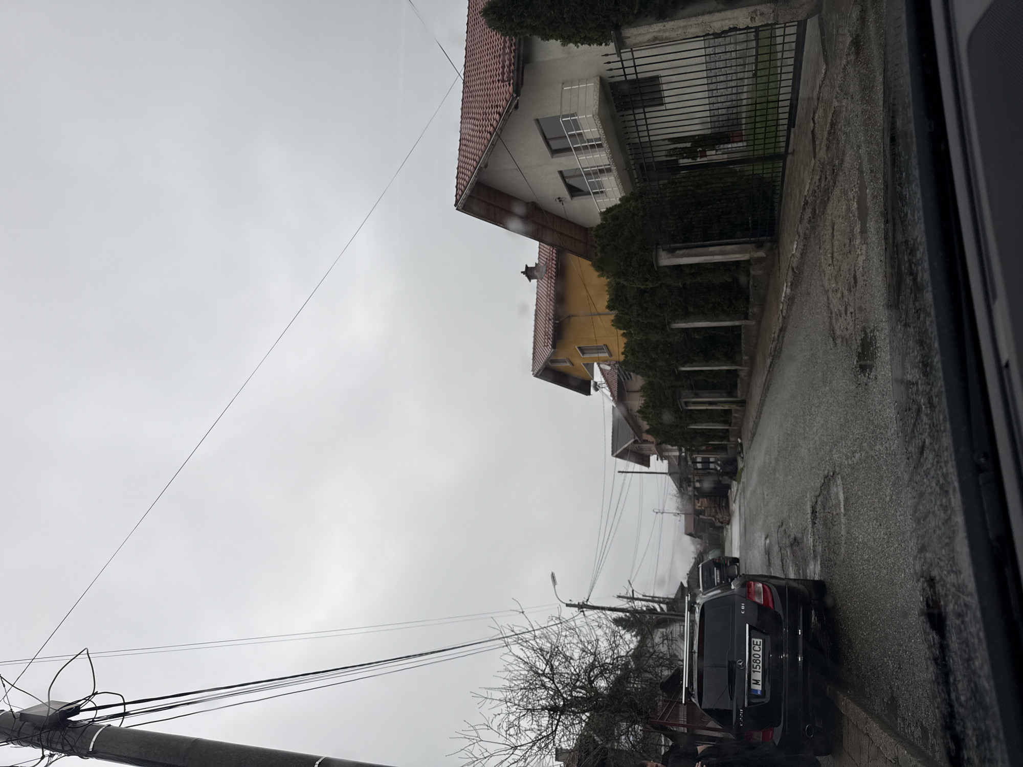 The image shows a residential street in Berkovitsa, Bulgaria on a cloudy day. The street is lined with houses, some with red tile roofs and others with metal fences. A black Mercedes C220 CDI is parked on the side of the road. Power lines are visible overhead. The road appears wet and patchy. The environment suggests a quiet, possibly older neighborhood within Berkovitsa.