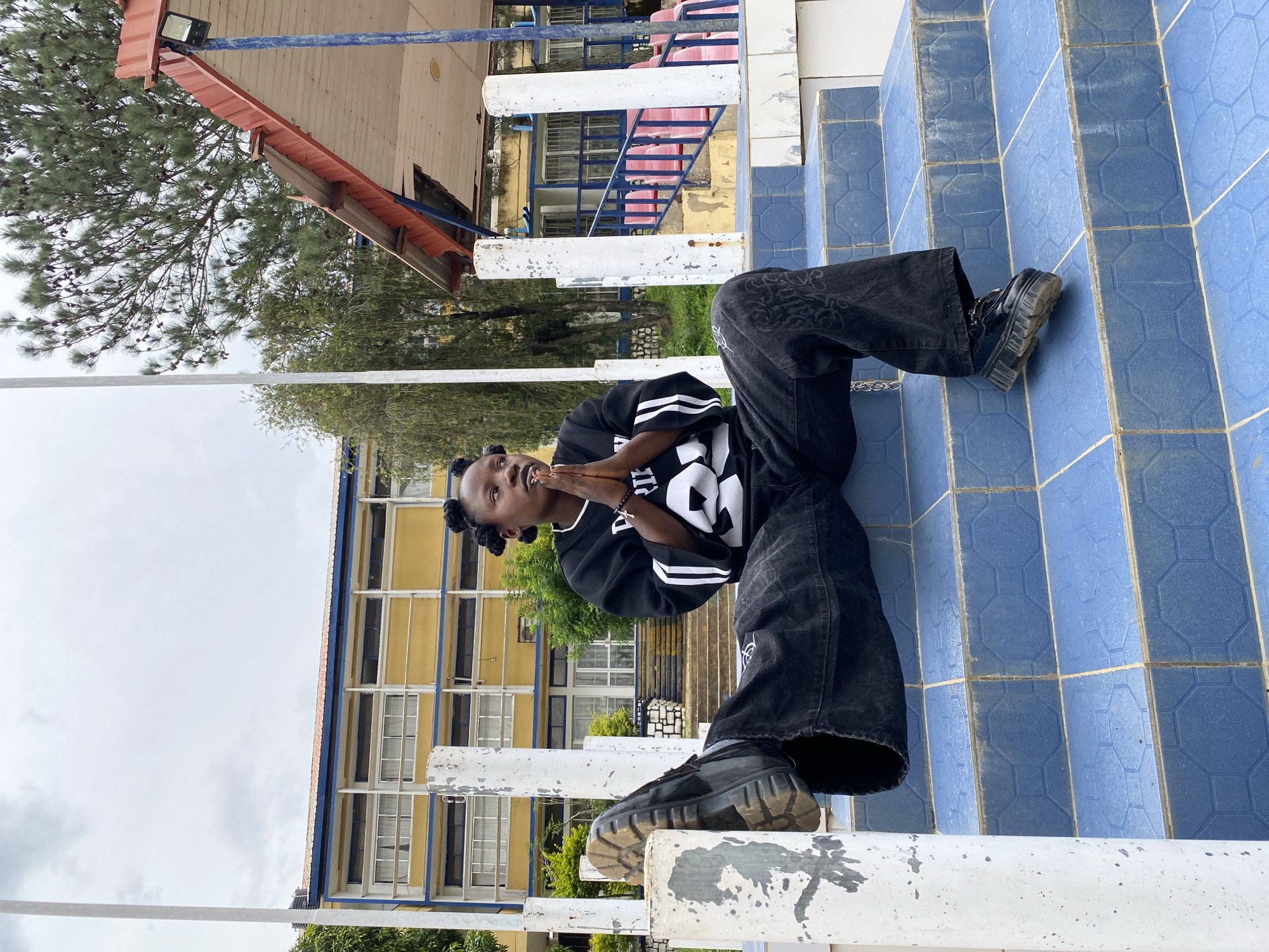 In Zawan, Nigeria, a young person with dark skin, styled hair in two buns, and wearing a black sweatshirt with white stripes and large black pants, is seated on blue tiled stairs outdoors. Their hands are clasped together in front of their chest. To their left, one leg is extended upwards. In the background, a yellow building with blue accents and many windows is visible, along with trees and a red-roofed structure. The scene is illuminated by overcast daylight.