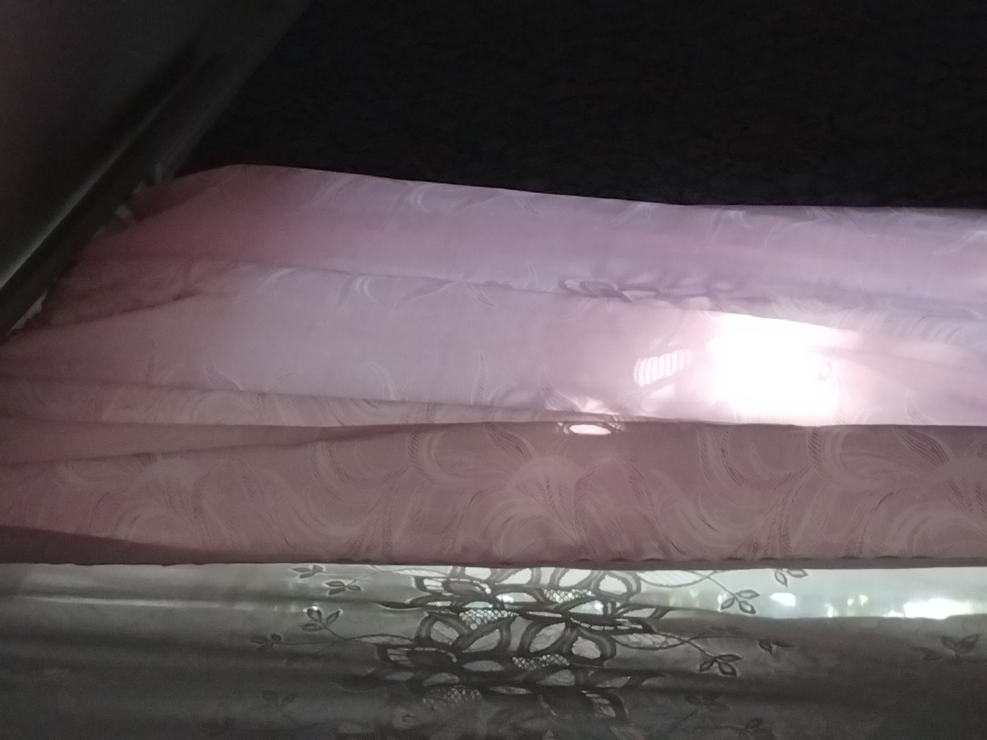 The image shows a close-up view of two draped curtains against a dark background. The left curtain is sheer and appears to have an embroidered floral pattern with leaves. The right curtain is a thicker, textured fabric with a swirling pattern, possibly in a light pink or lavender hue. Sunlight is filtering through a gap in the curtains, creating a bright, blown-out area in the center of the image, illuminating the texture of the right curtain. The overall scene suggests an indoor setting, likely a window with curtains. There are no people, animals, or any discernible activity depicted. The context of Pervozvanivka, Ukraine, cannot be determined from the visual information presented in the image.