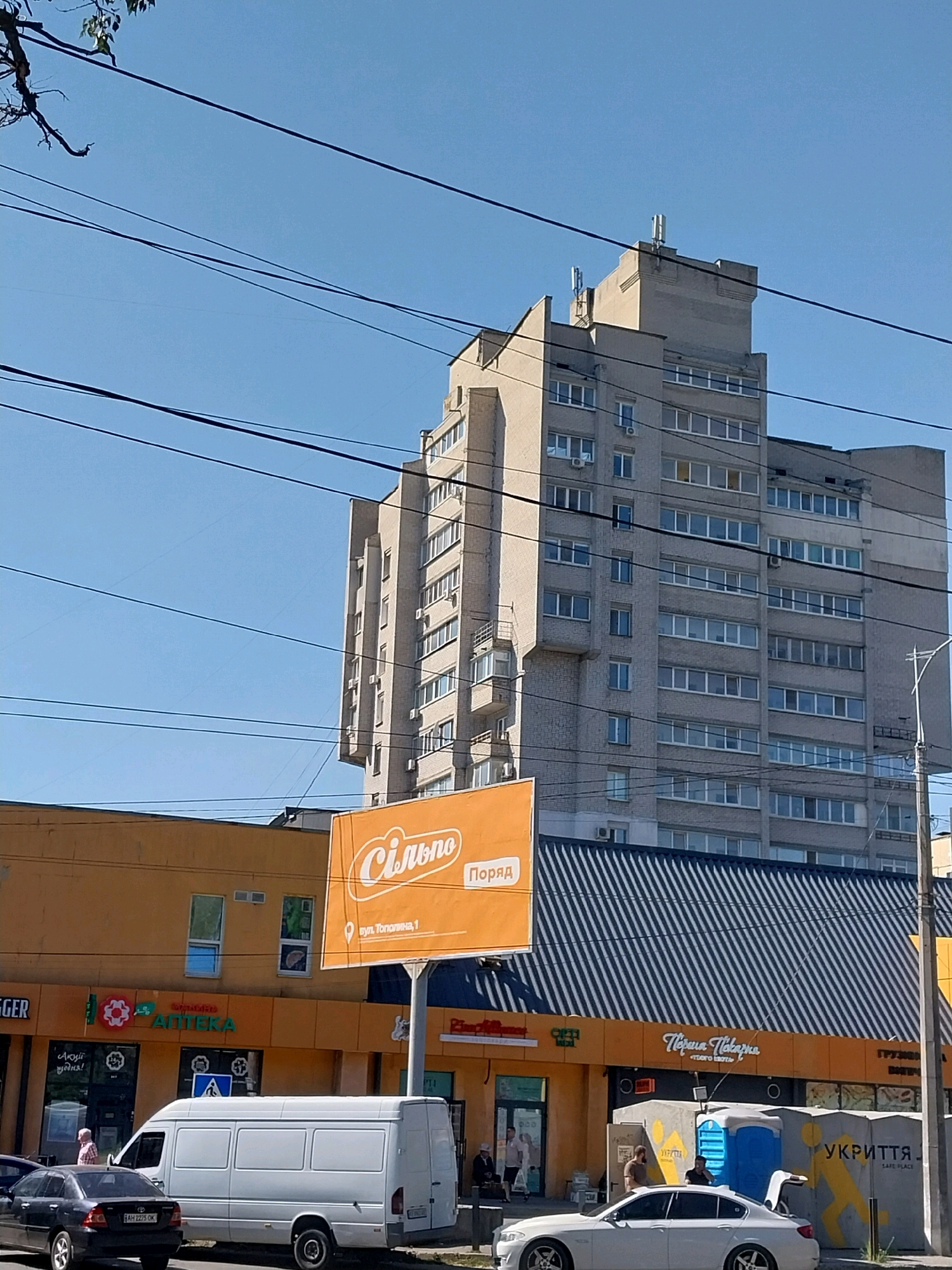 The image shows a street scene in Dnipro, Ukraine, under a clear blue sky with several overhead wires crisscrossing the frame. In the foreground, a black sedan is parked on the left, and a white van is parked in the center. To the right of the van, a white sedan is parked. Behind the vehicles, a row of commercial buildings is visible, primarily in orange and dark blue. One building has an orange awning with the text "Сільпо" and "ПОРЯД" on a large billboard. Another building has the text "АПТЕКА" with a logo. Further to the right, another storefront has the text "Пекарня" and "УКРИТТЯ". People are sparsely visible walking in front of the businesses. Dominating the background is a tall, multi-story concrete building with many windows.