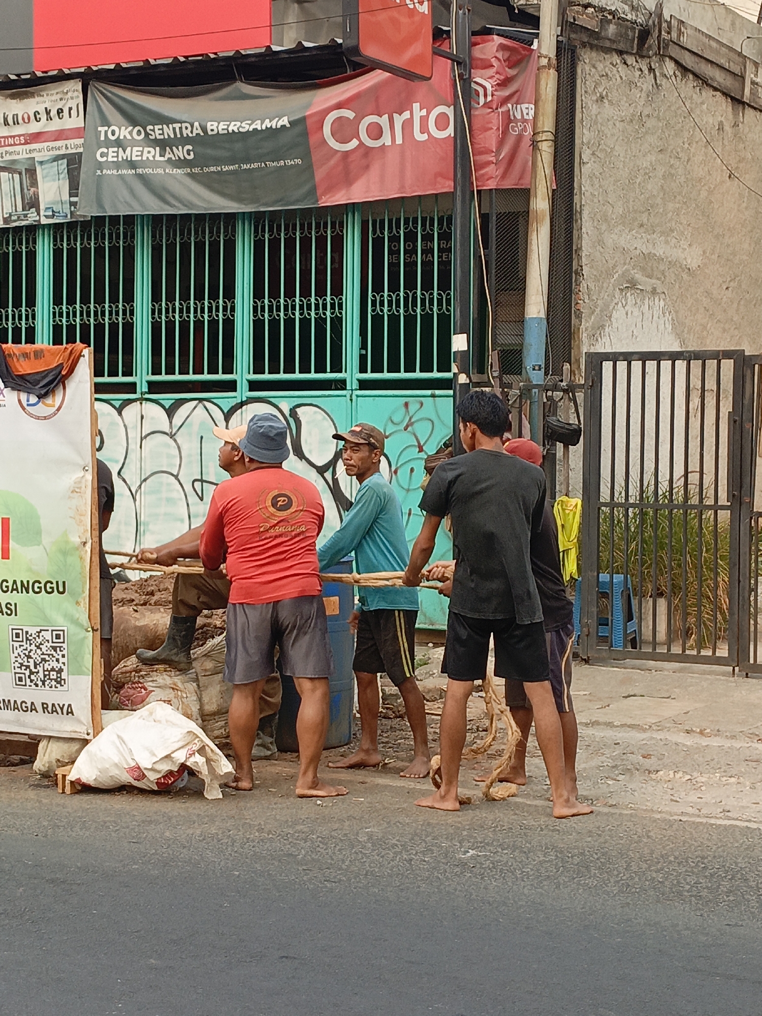 The image depicts a street scene in East Jakarta, Indonesia, likely in a commercial or semi-industrial area. Four men are actively engaged in pulling on wooden poles, suggesting they are moving a heavy object, perhaps a large bundle or a vehicle part, that is partially obscured by a white tarp and burlap sacks on the left side of the frame. The men are dressed casually, some in t-shirts and shorts, and appear to be working together. One man wears a blue hat, another a beige cap.
The background features a building with a prominent banner that reads "TOKO SENTRA BERSAMA CEMERLANG" and lists an address: "JL. PAHLWAN REVOLUSI, KELENDER, KEC. DUREN SAWIT, JAKARTA TIMUR 13470". This banner indicates the specific location within East Jakarta. The building has teal-colored metal gates with ornate detailing, some of which are covered in graffiti. Adjacent to the building is a utilitarian structure with a rough, textured wall and a metal fence. A blue plastic stool and a yellow cloth are visible near the fence. The ground is paved with asphalt and gravel. The lighting suggests it is daytime, possibly late afternoon, given the warm tones and shadows. The overall atmosphere is one of manual labor and everyday urban activity.