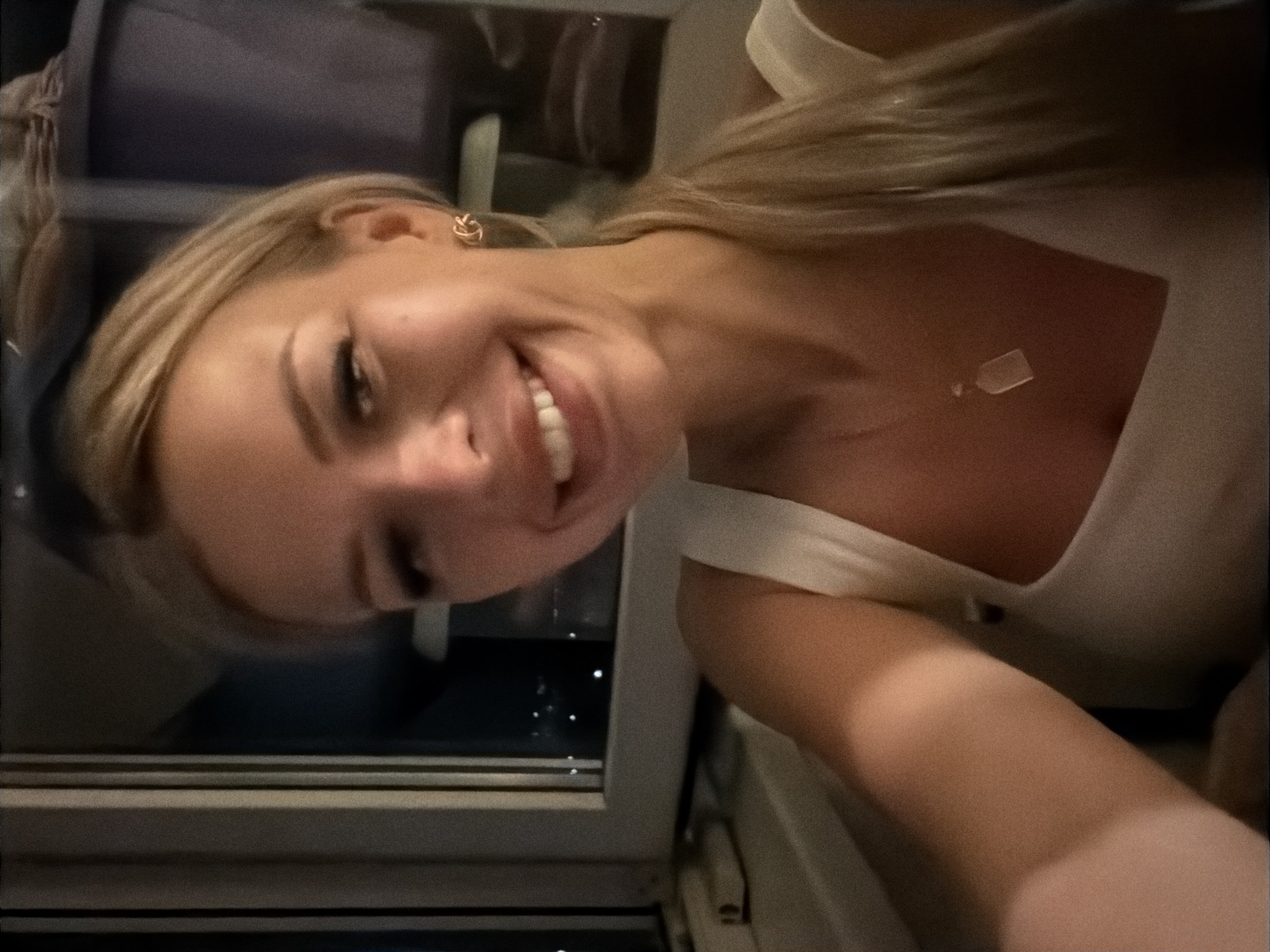 In an indoor selfie, a fair-skinned blonde woman with light makeup smiles directly at the camera. She is wearing a white top with thin straps and a light-colored pendant necklace. A small, gold earring with an abstract design is visible on her right ear. Her blonde hair is pulled back into a ponytail. Behind her, a window reveals a blurred city skyline at night, with small lights indicating buildings and distant urban activity. The window frame appears to be dark and possibly made of metal. The overall lighting is dim, with a warm cast, suggesting an evening or indoor setting.