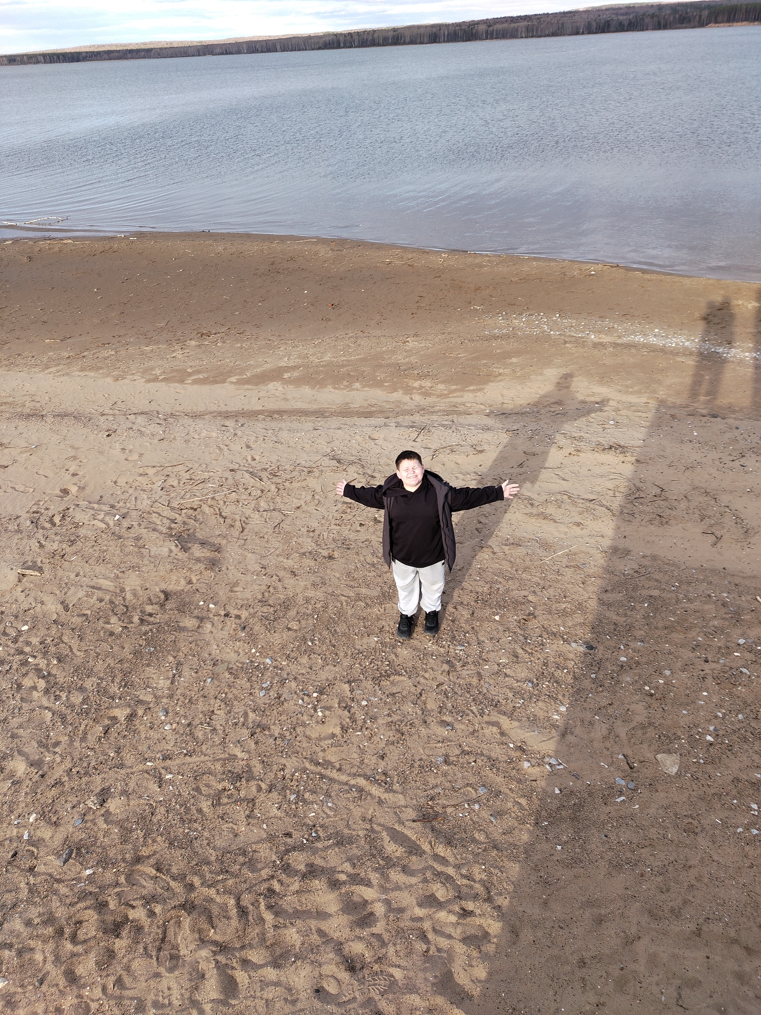 A person stands on a beach with arms outstretched, facing the camera. The beach is sandy and extends from the lower half of the frame to a transition zone of darker sand nearer the water's edge. Beyond the beach is a body of water, presumably a lake or bay, stretching toward a distant shoreline lined with trees. The sky is overcast. The person is wearing a black shirt, light-colored pants, and a dark jacket. The shadow of the photographer is visible to the right. The location is Unknown, Unknown.