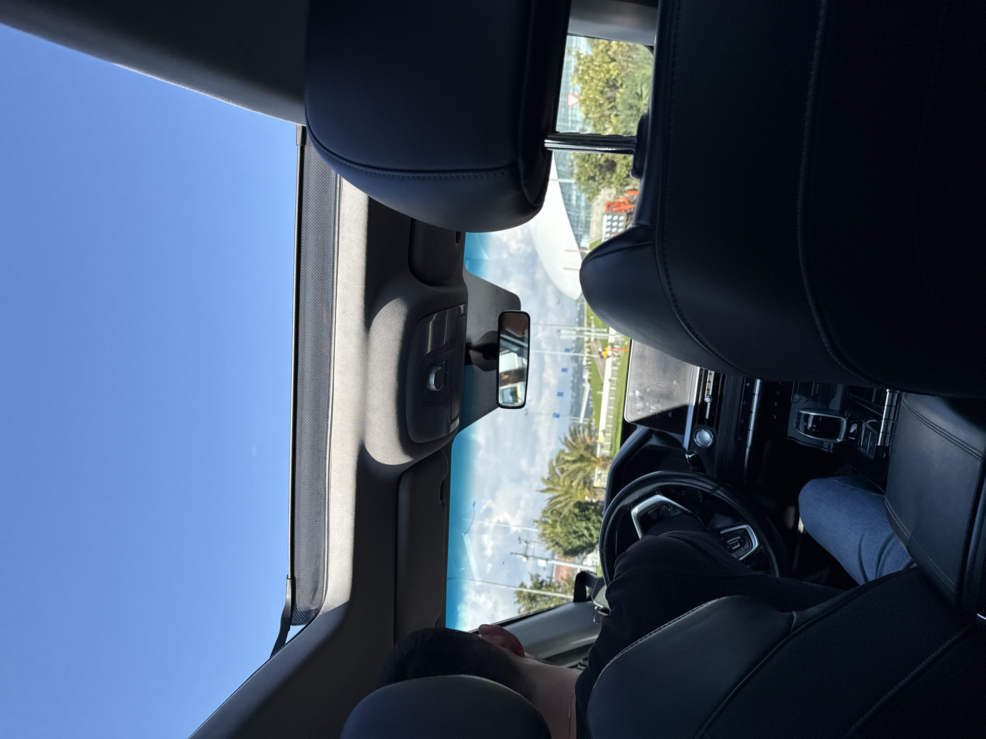 The image, taken from inside a modern car, offers a view of the interior and the outside environment through the windshield and panoramic sunroof. The perspective is from a rear seat, looking forward.
In the foreground, the tops of two black leather front seats are prominent. The driver is visible from behind, with their dark hair, head, and shoulders rising above the left seat's headrest. They are dressed in a dark-colored top, with blue jeans partially visible on their lap. A second person's leg, also clad in blue jeans, is partially seen towards the bottom right, suggesting a front passenger. The car's interior features include a black dashboard, a partially visible steering wheel with metallic accents, and a rearview mirror reflecting a cloudy sky.
Beyond the car's interior, the outdoor scene is bathed in bright daylight under a clear blue sky, dotted with some scattered white clouds. Green palm trees are distinct elements of the landscape. In the distance, several modern structures are visible, including a prominent large white building with a curved design. Other outdoor elements include utility poles, power lines, and a triangular traffic sign with a red border.
The time of day is clearly daytime, characterized by bright sunlight and clear weather. The presence of palm trees and contemporary architecture aligns with the specified location context of Sirius, Russia, known for its coastal setting and modern developments. No legible text is visible within the image.