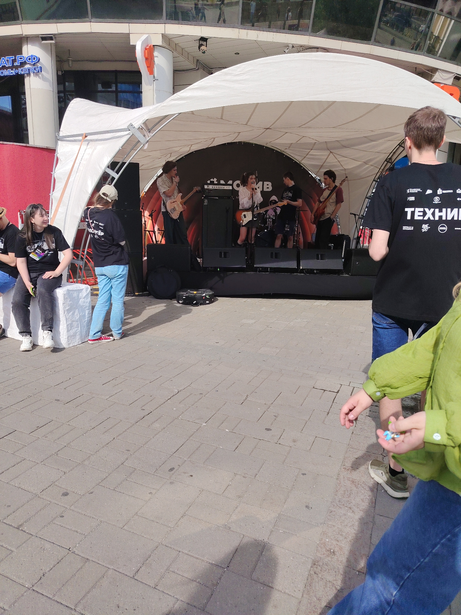 In Yekaterinburg, Russia, a live music performance is taking place outdoors on a temporary stage. A band, comprising at least three males and one female, is performing on a raised platform covered by a white tent. The musicians are playing guitars and there is a drum set visible. One of the male musicians is singing into a microphone.
In the foreground, on the left side of the image, two young women are seated on white cubes. The woman on the left is wearing a black t-shirt and jeans and is looking towards the stage. The woman on the right is wearing a black t-shirt, a light-colored cap, and light blue jeans, and is also looking towards the stage.
On the right side of the image, a person wearing a black t-shirt with white text and blue shorts is standing, facing away from the camera. Another person, partially visible, is wearing a light green shirt and blue jeans, and is holding a small blue object in their hand.
The stage area is set up on a paved pedestrian area, likely a plaza or a street closed for the event. In the background, a building with glass windows is visible, reflecting the sky and surrounding environment. The overall atmosphere suggests a public event or festival.