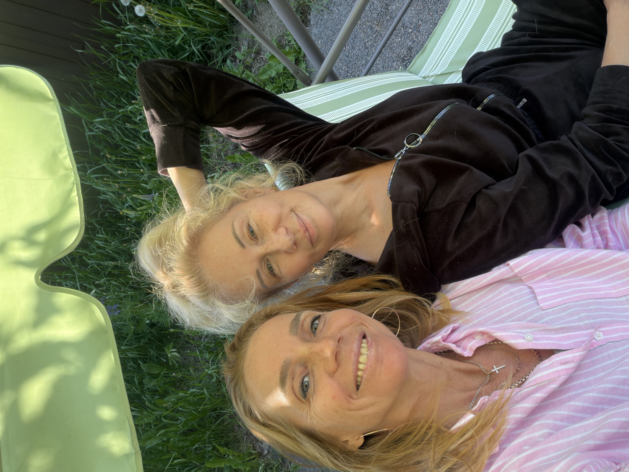 Two blonde women are depicted lying down outdoors, looking up at the camera with relaxed, smiling expressions. The woman positioned higher in the frame has lighter, curly blonde hair and is dressed in a black long-sleeved top with a prominent zipper. She is reclining on an outdoor lounge chair with green and white stripes. The second woman, positioned below, has golden blonde hair and is wearing a pink and white striped collared shirt. She accessorizes with a silver chain necklace featuring a small cross pendant and large hoop earrings.
The setting appears to be a sunny outdoor space, likely a garden or backyard in Steklyannyi, Russia. Lush green grass is visible around the women, along with hints of other outdoor elements like a solid green mat or chair on the left. The bright, natural daylight suggests it is daytime, casting clear but soft illumination on their faces. The overall mood conveyed is one of contentment and leisure, with both women appearing to enjoy the sunny outdoor environment. No visible text is present in the image.