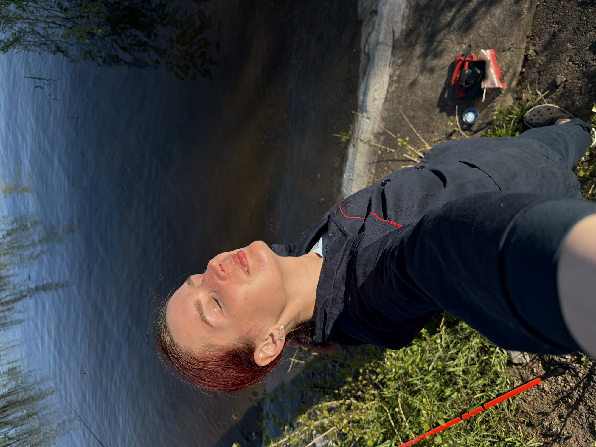 A person with red hair and fair skin is taking a selfie near a body of water, possibly a river or lake. They are wearing a dark jacket with red trim and dark pants, along with what appears to be a Croc shoe. A backpack and other personal items are on the ground. A red and black cane is visible near the grass. The location appears to be outdoors, possibly in a park or near the waterfront, with a concrete embankment and grassy area visible. Given the user input, this location is assumed to be somewhere in Chaikovsky, Russia.