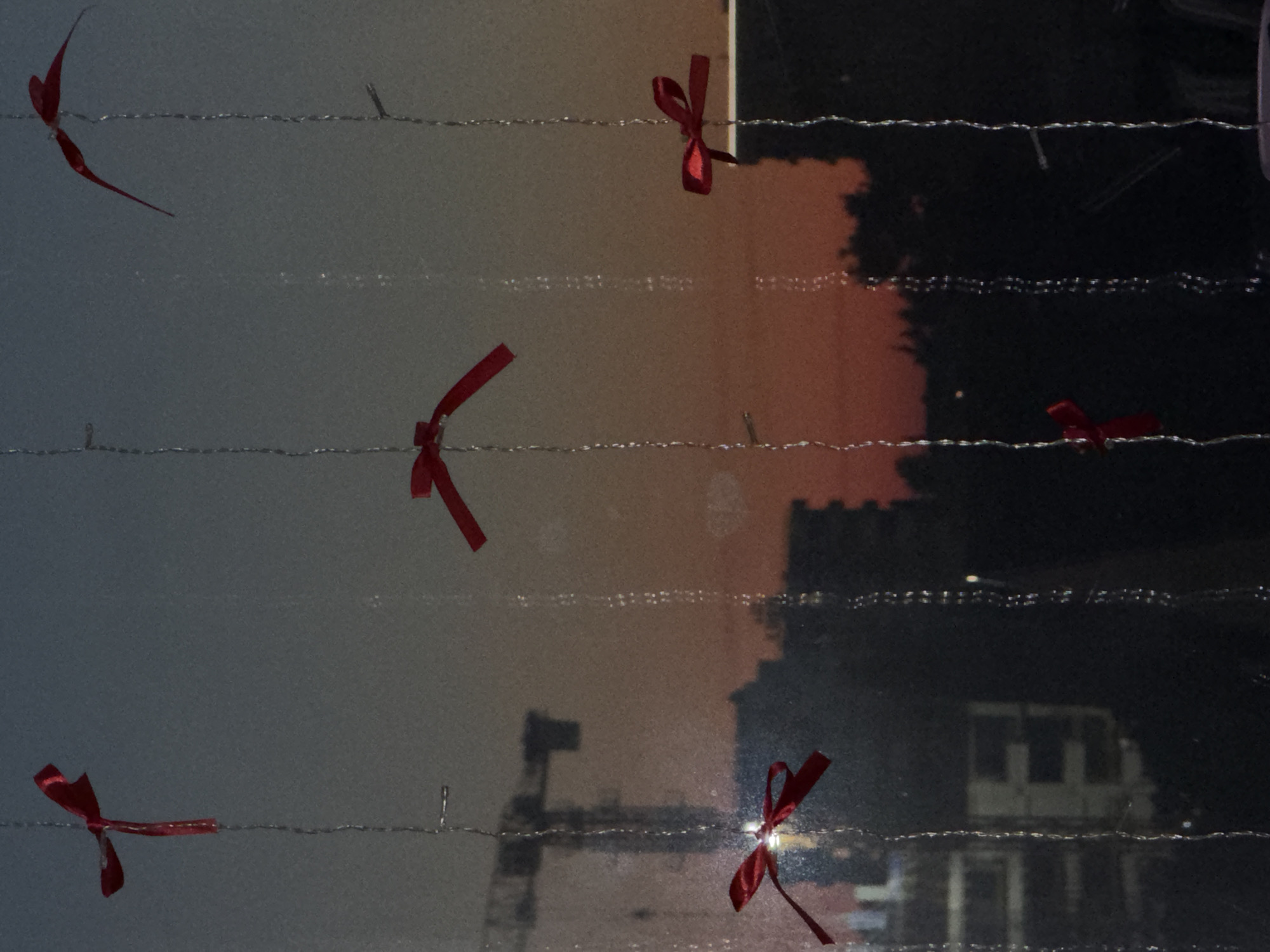 A dark, grainy image shows a twilight view through a window with water droplets. Several red ribbons are tied to thin, silver strings, hanging vertically. In the background, the silhouettes of buildings and a construction crane are visible against a gradient sky of orange, pink, and blue. The scene appears to be an urban setting. There is no information in the image to indicate it is located in Sirius, Russia.