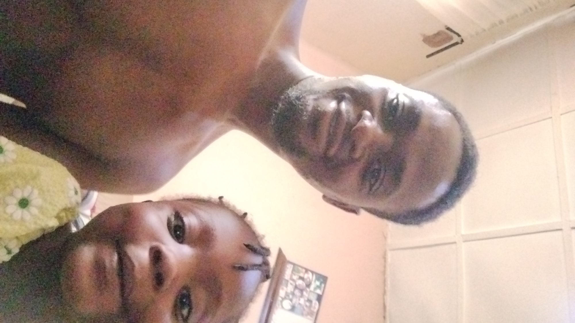 The image depicts two individuals: an adult male with a beard and a child. They are positioned with their faces close to the camera, suggesting they are taking a selfie. The adult male is shirtless. Behind them, the background includes a wall painted in a light color and a white tiled wall. A framed photograph is partially visible. The exact location in the city of Jos, Nigeria, cannot be determined from the visual information provided.