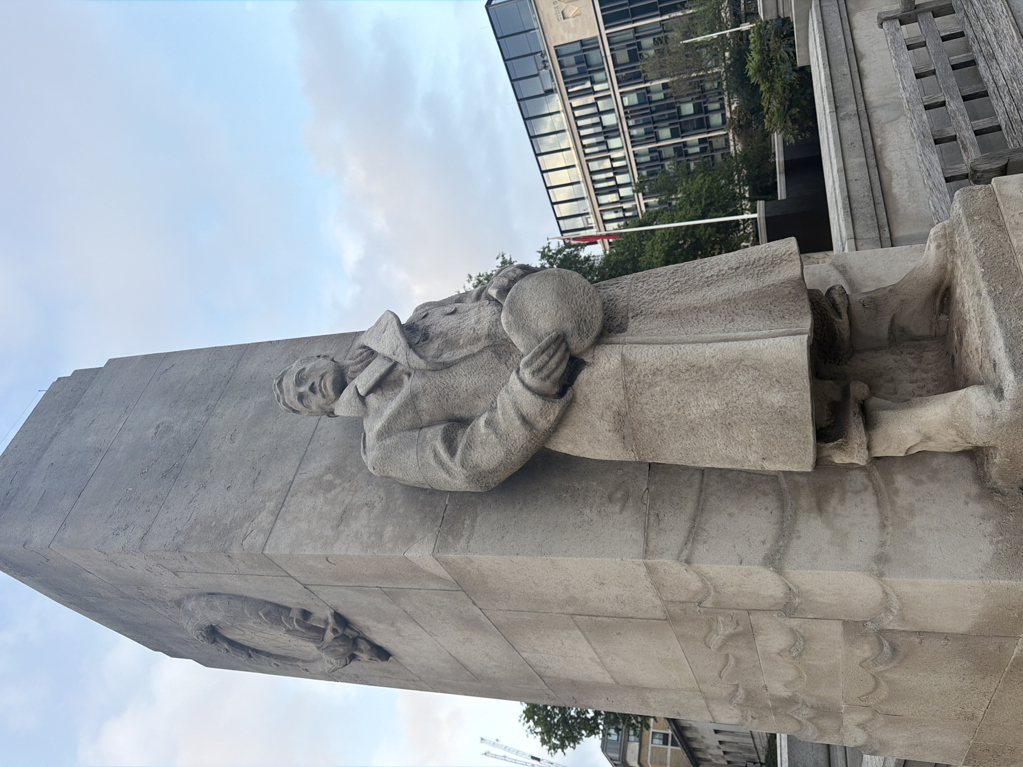 The image depicts the monument to the Merchant Navy in London, United Kingdom. The monument features a sculpted figure of a merchant seaman holding a globe, standing atop a tall stone pillar. The pillar is adorned with decorative carvings. Behind the monument, modern buildings are visible, including one with mirrored glass windows. Trees and flags can also be seen in the background. The sky is overcast. The photo is taken at a low angle, looking up at the monument.