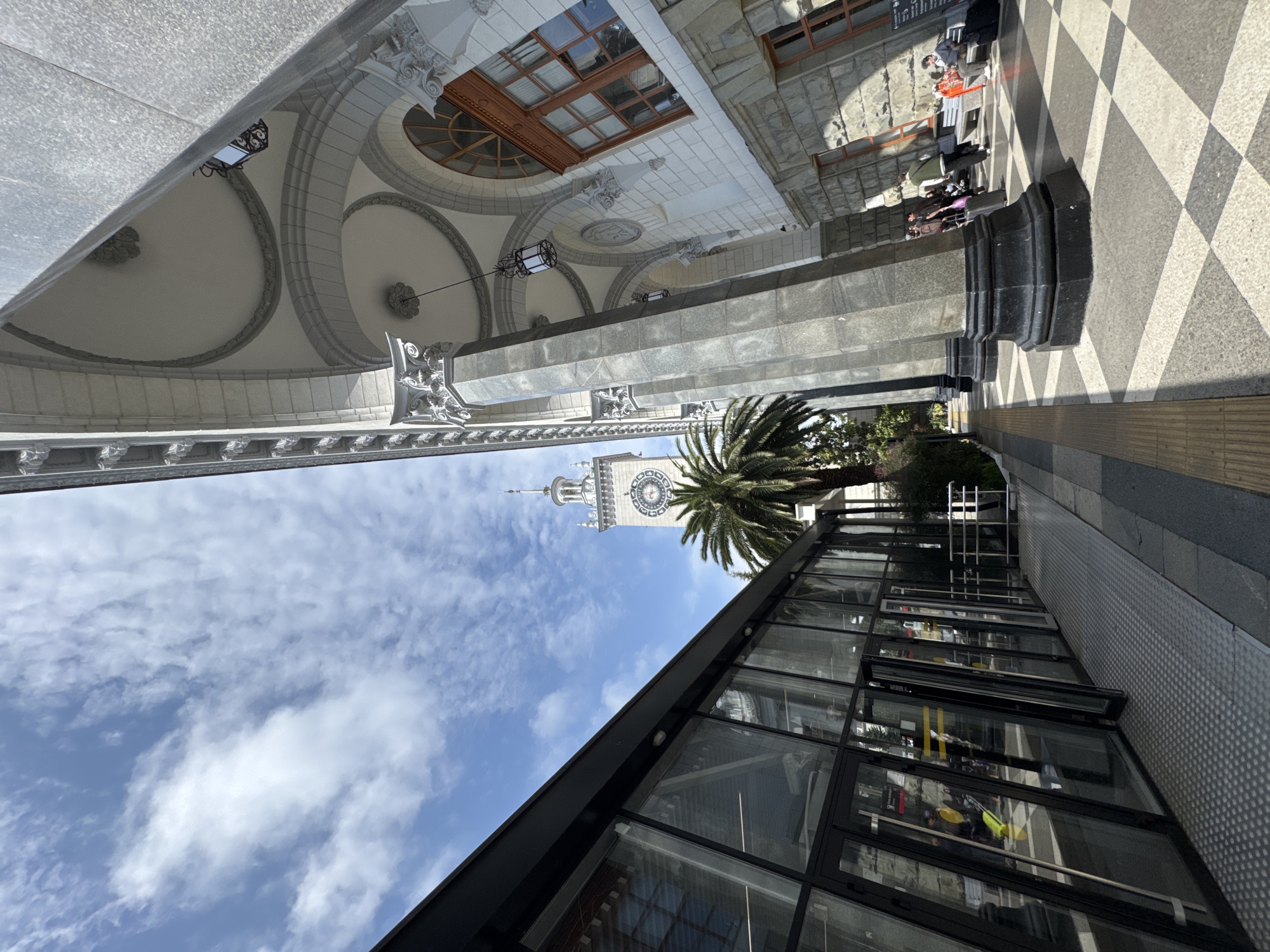 The image depicts a low-angle view in Sochi, Russia. The location is near the Sochi Railway Station, with the station's clock tower visible in the background. A palm tree partially obscures the clock tower. A modern glass-walled structure is in the foreground. Above the ground level, a classical building with arched ceilings, decorative moldings, and windows is shown. The ground is paved with a black and white checkered pattern. People are seated at tables on the checkered pavement, suggesting an outdoor cafe or seating area. There are no interactions between people, but there are stationary people.