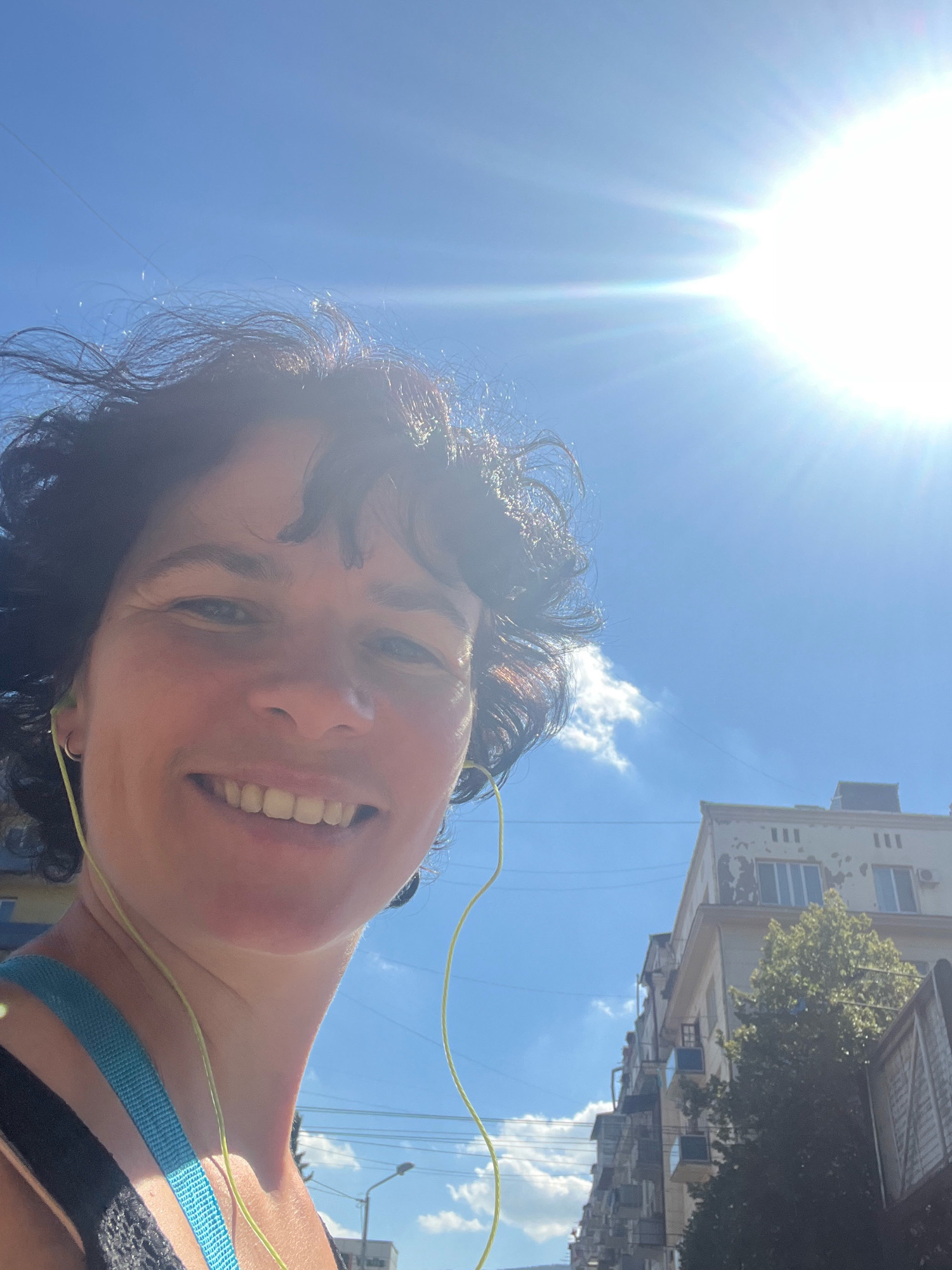 A close-up, low-angle selfie is taken outdoors on a bright, sunny day, with the sun prominently visible in the upper right quadrant, casting lens flare. The subject, a woman with dark, curly hair and fair skin, is smiling and looking directly at the camera. She is wearing earphones, with one earbud visible in her left ear, and a turquoise strap is visible over her left shoulder, suggesting athletic wear. The background features a clear blue sky with scattered white clouds, and beyond that, buildings are visible, some appearing weathered. One building on the right has a light-colored facade with multiple windows and what appear to be architectural details like pilasters. The scene suggests an urban environment, possibly in Tbilisi, Georgia, given the architectural style.