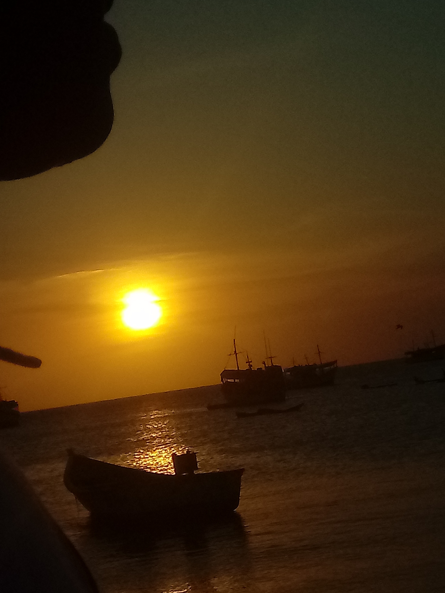 The photograph captures a sunset over the sea in Juan Griego, Venezuela. Multiple boats are silhouetted against the bright orange and yellow sky. The sun is positioned high in the sky, reflecting brightly on the water's surface. The water is calm, with gentle ripples reflecting the light. The upper portion of the image is partially obscured by a dark shape on the left side, presumably a person or object close to the camera. The horizon line is visible, separating the sea from the vibrant sky. A bird is seen flying in the distance.