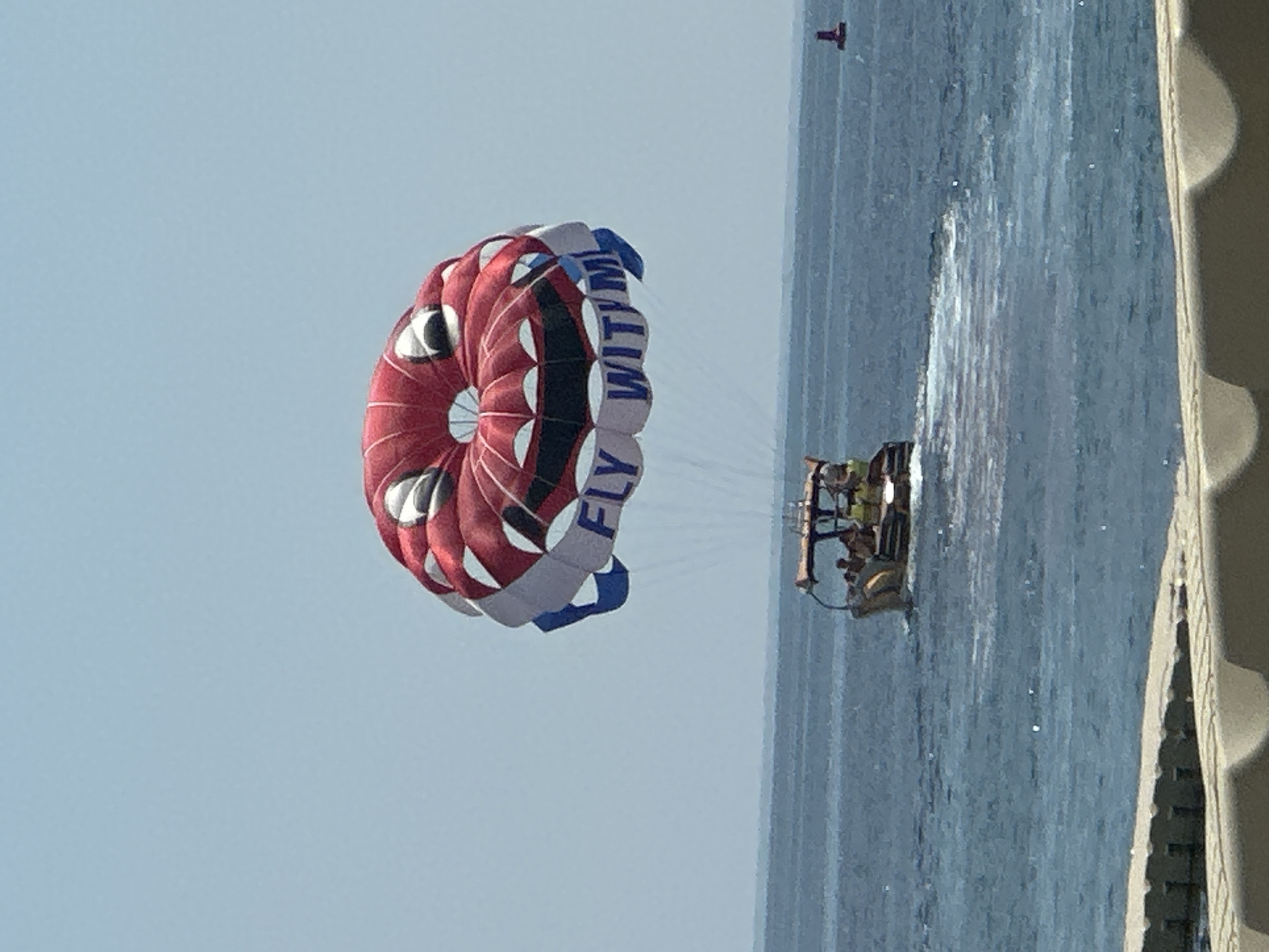 The image shows a bright, sunny day with a clear blue sky over the sea. In the foreground, there is a concrete structure, possibly a pier or walkway. In the middle ground, a boat is moving across the water, leaving a white wake. The boat appears to have several people on board. In the background, a parasail shaped like a large, smiling face is being towed behind the boat. The parasail is red and white with blue accents and has the words "FLY WITH ME" written on it. A small red buoy is visible in the distance to the right of the boat. The overall scene depicts a recreational activity taking place on the water, likely a tourist attraction in Budva, Montenegro, given the coastal setting and the presence of leisure activities.