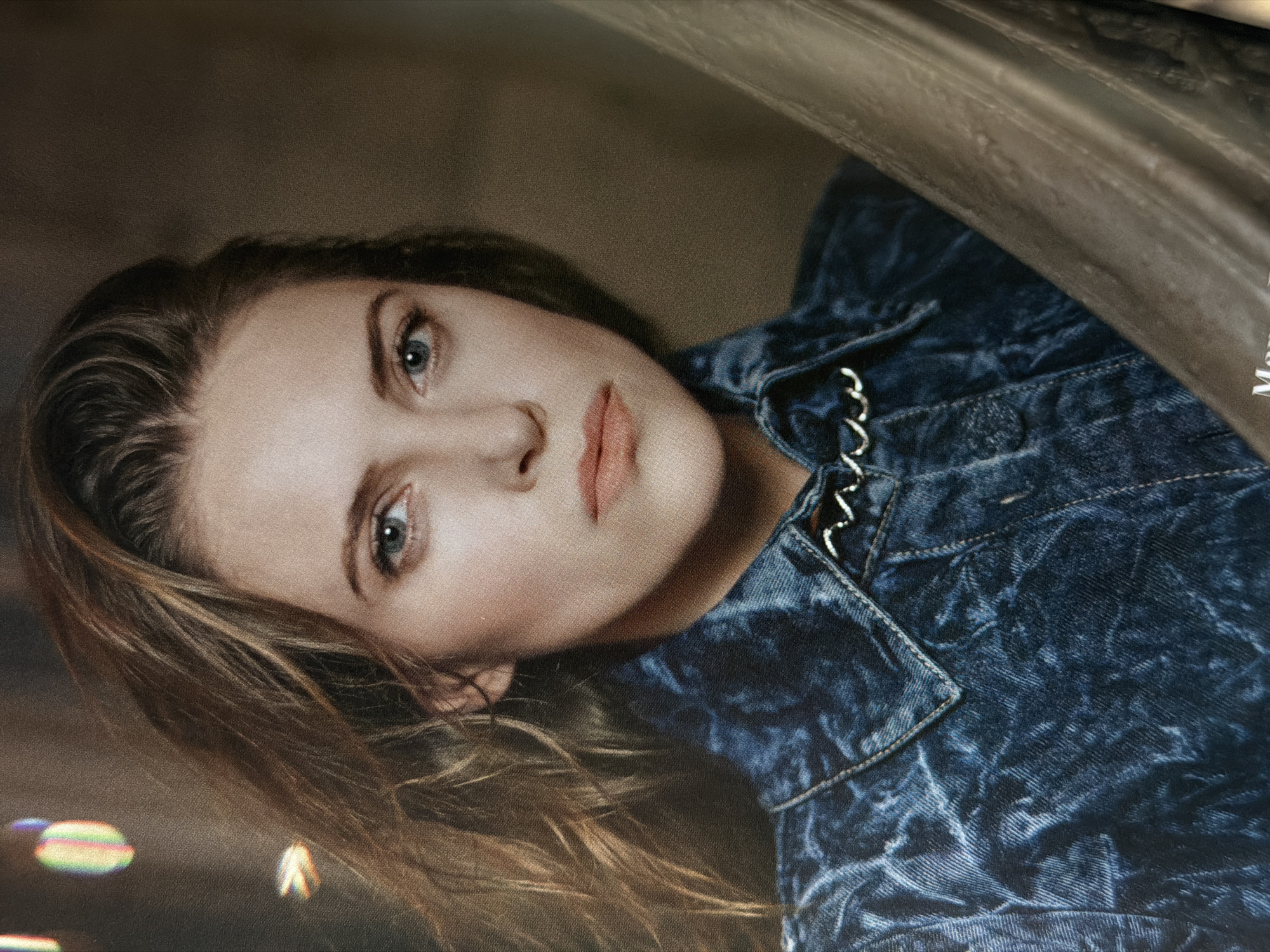 A medium-close, slightly angled shot shows a woman with fair skin and light blue eyes. She is looking upwards and slightly to her left. Her hair is light brown and styled with loose waves. She wears a dark blue denim jacket with silver chain detailing at the collar. The background is blurred, with bokeh lights suggesting an urban environment. A dark grey, possibly leather, surface is visible in the upper right corner. The location, Moscow, Russia is not discernible in this image.