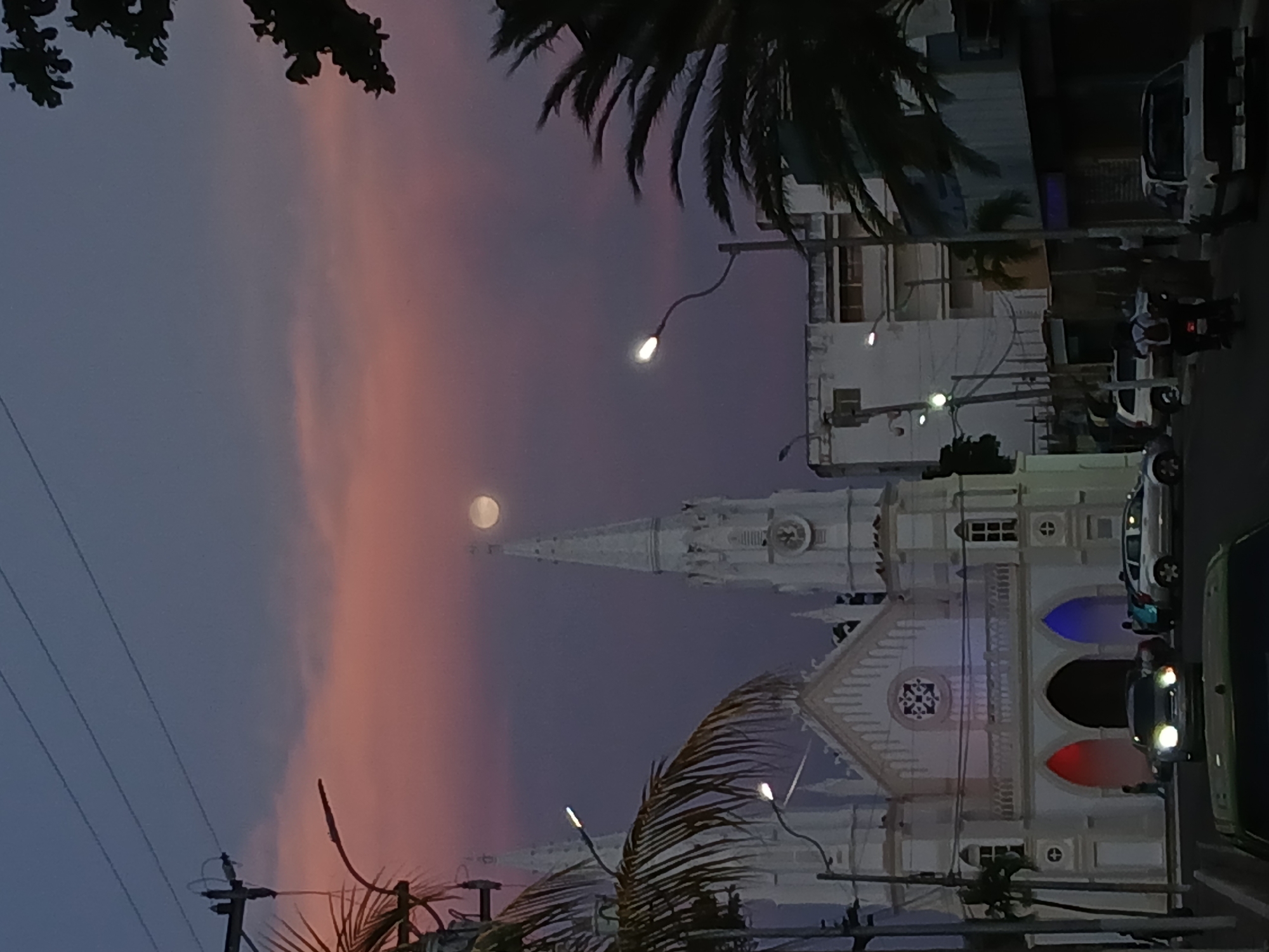 This image captures a serene evening street scene in Juan Griego, Venezuela, bathed in the soft glow of dusk or twilight. The sky transitions dramatically from a deep purplish-blue to a gentle, cloudy pink-orange, creating a picturesque backdrop.
The central subject is a grand, white church with intricate architectural details, including multiple spires and towers. A prominent, tall spire points towards the sky, with a clock face visible near its apex. The church's facade features arched doorways, some of which are illuminated with distinct colored lights – red and blue – adding a vibrant touch to the structure.
High in the sky, a bright, full or near-full moon shines, positioned slightly behind the church's main spire, enhancing the tranquil atmosphere. Dark silhouettes of palm fronds frame the scene, suggesting a tropical or coastal environment, consistent with the location context.
On the right side, a street runs alongside the church, populated by several vehicles. Cars are visible with their headlights on, and a white van or truck is parked further down. Streetlights cast their warm glow, contributing to the evening illumination. Utility poles and power lines crisscross the upper left portion of the frame. A few blurry figures are discernible near the church entrance and vehicles, indicating the presence of people, but no specific activity or event beyond typical street life is clearly depicted. The overall impression is one of peaceful beauty during the transition from day to night.