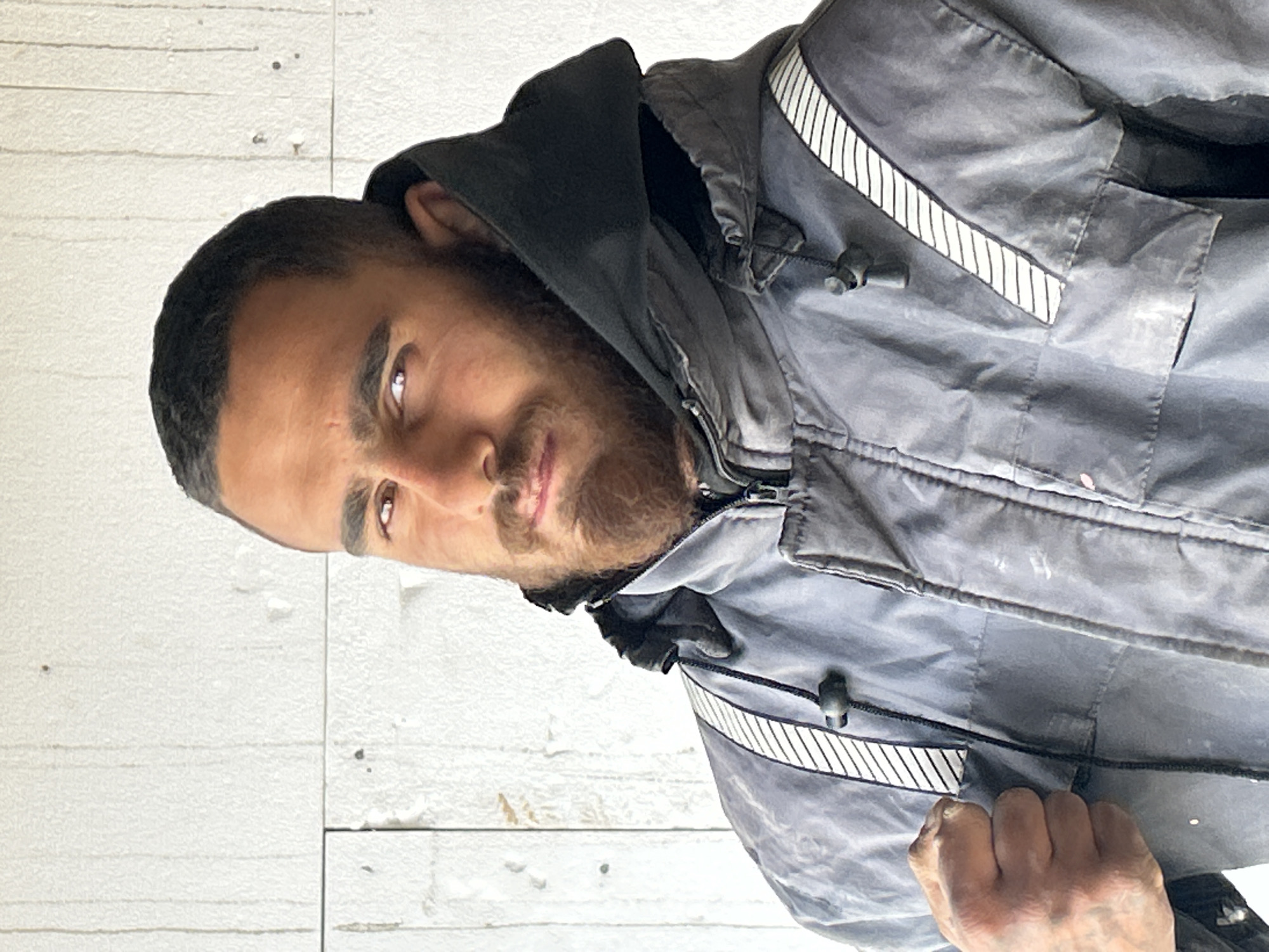 A man with short, dark hair, a dark beard, and brown eyes is facing the camera. He is wearing a gray hooded jacket with white striped accents on the shoulders. He is positioned in front of a white wall, possibly textured or made of tiles. The image does not contain enough information to determine the location in Tsarevo, Bulgaria.
