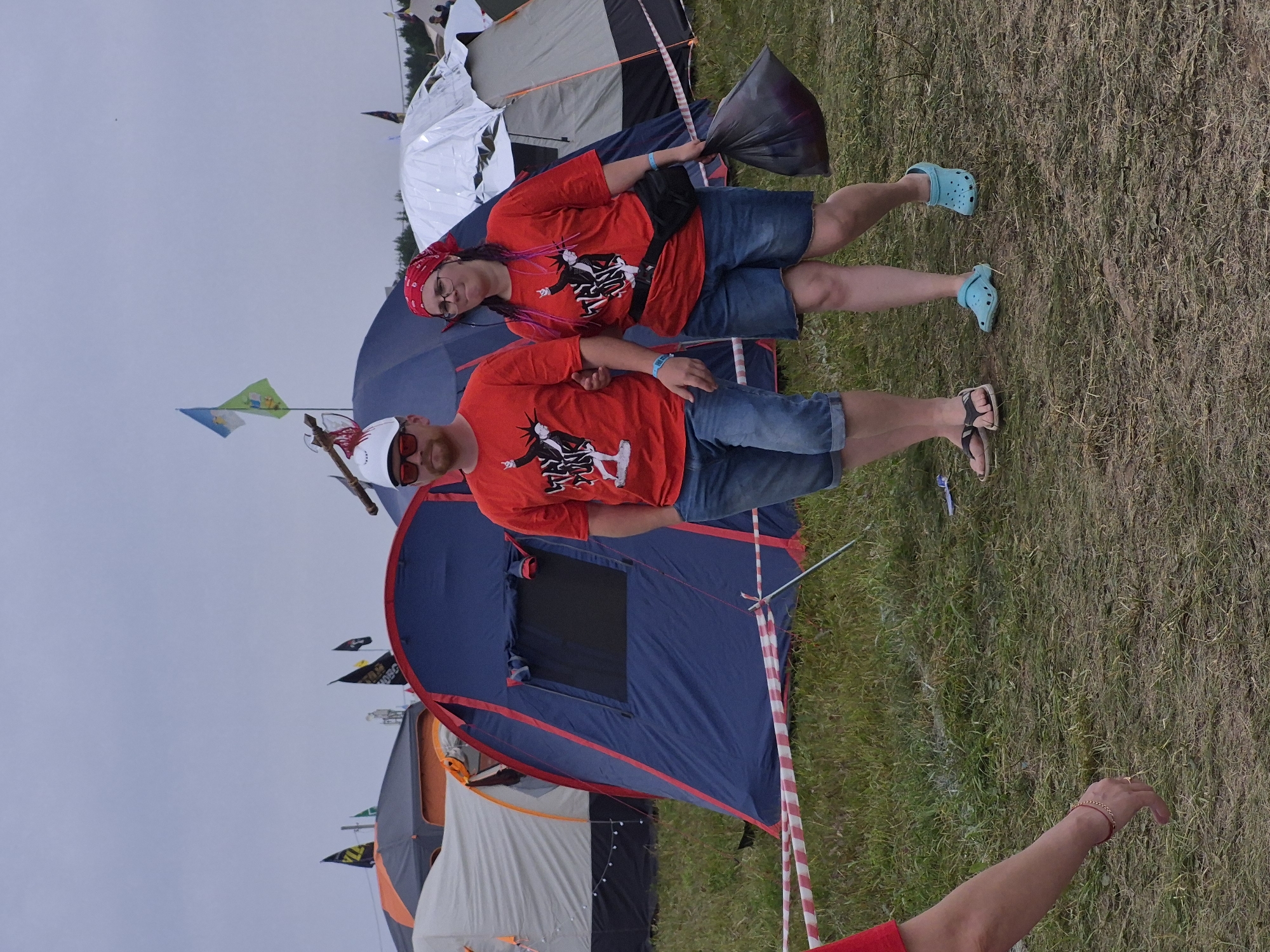 In Lagunovo, Russia, a man and a woman are walking arm-in-arm in a grassy outdoor area, possibly a festival campsite. They are both wearing red t-shirts with a graphic print. The man has a white cap and sunglasses, and is wearing denim shorts. The woman has a red bandana on her head, glasses, and is wearing denim shorts. She is carrying a black plastic bag. They are walking past several tents of various colors, including a blue and red one, and a white one covered in foil. Red and white striped tape is visible on the ground in front of them, marking a boundary. The sky is overcast.