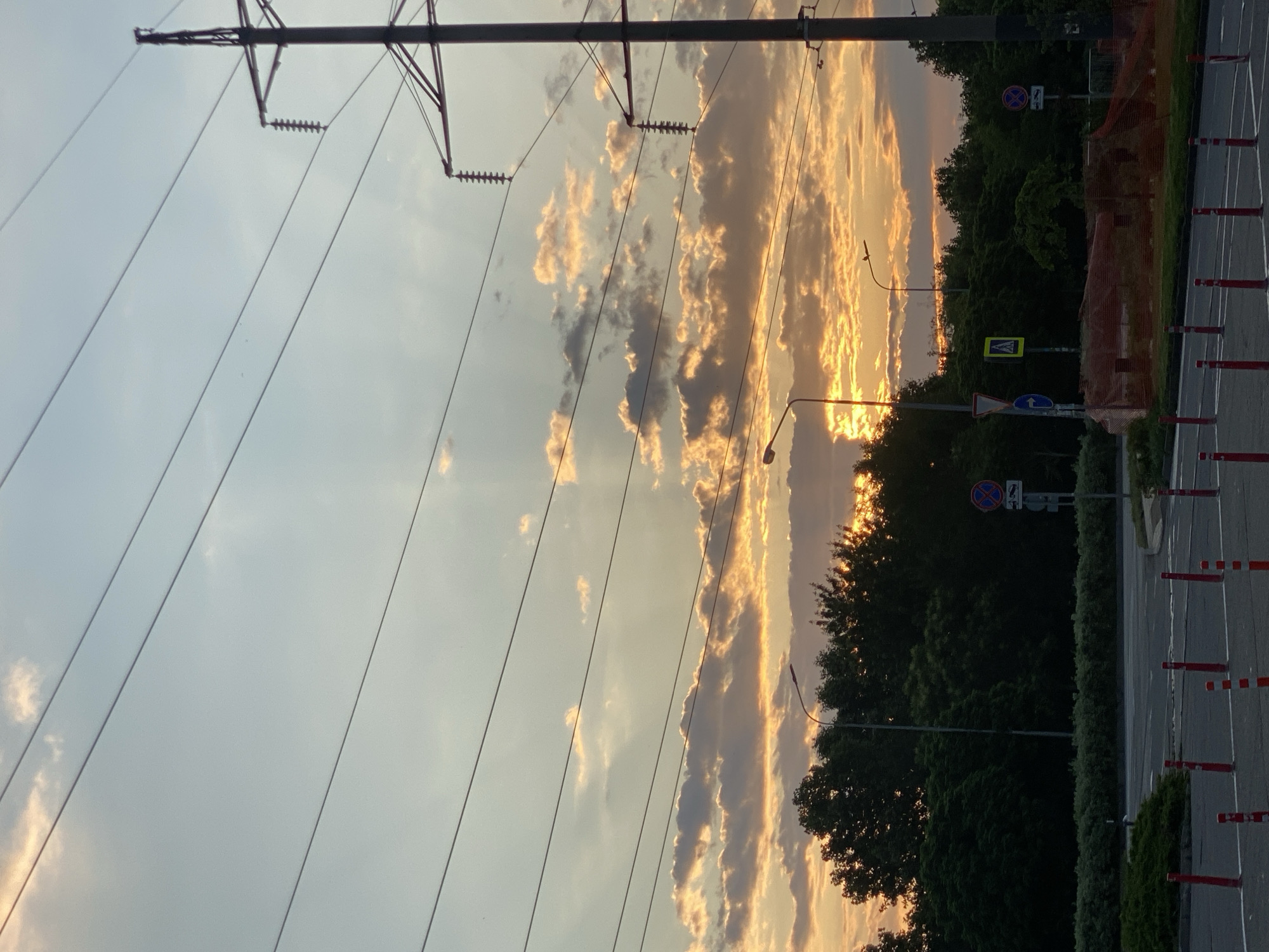 The image depicts an outdoor scene at dusk, likely on the outskirts of a city. The sky is filled with dramatic clouds, illuminated by the setting sun with warm orange and yellow hues. Several power lines stretch across the frame, crisscrossing the sky. On the right side, a tall electrical pylon stands prominently. In the middle ground, a dense line of trees forms a dark silhouette against the bright sky. A street light is visible amongst the trees. The foreground shows a paved road with lane markings and red and white bollards, suggesting it is a street or parking area. Various traffic signs are also present, including a pedestrian crossing sign and a "no stopping" sign. The overall atmosphere is serene, with the natural beauty of the sunset contrasting with the man-made infrastructure.