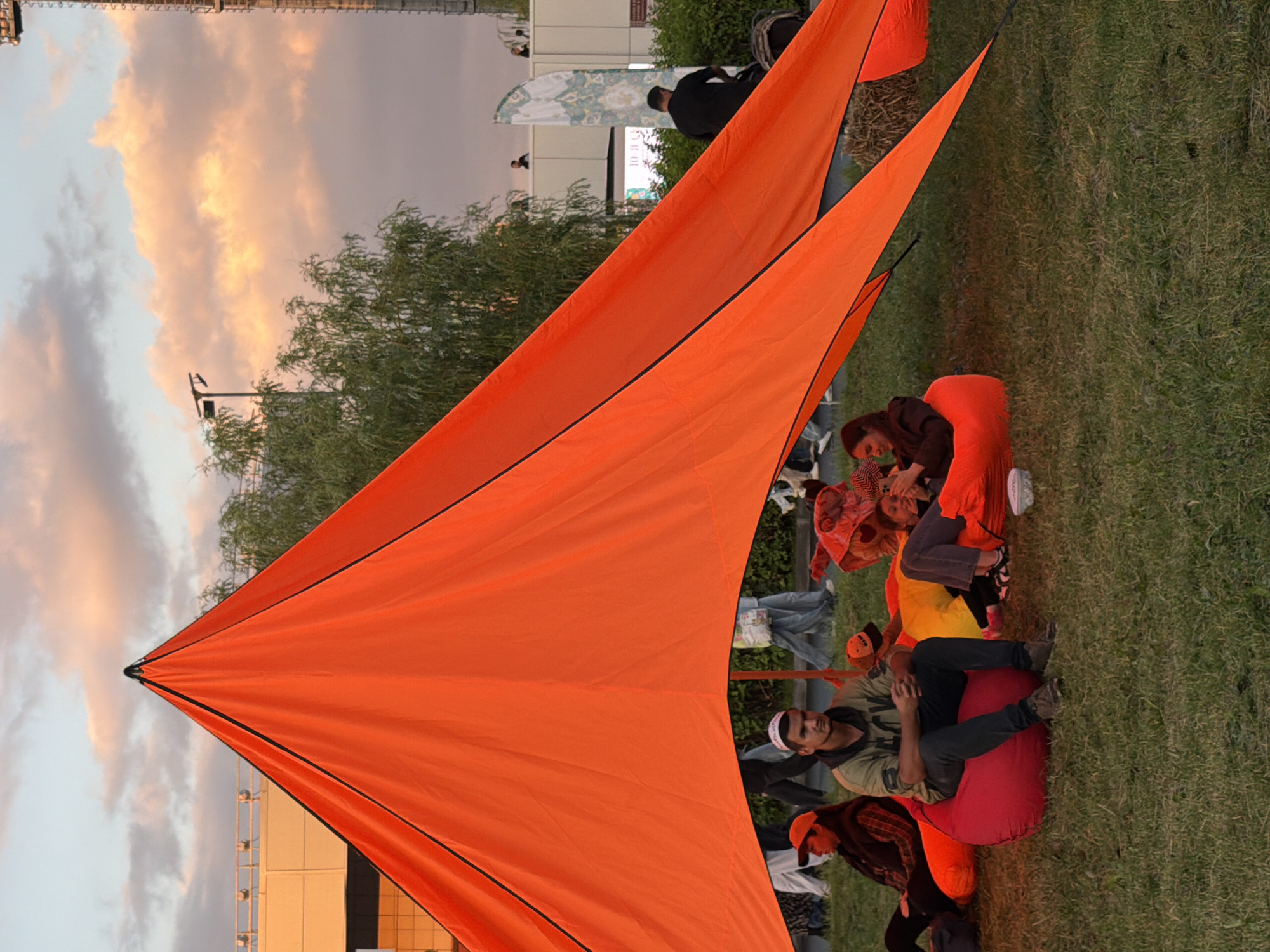 The image depicts a group of people relaxing on beanbag chairs under a large, orange tent structure. The setting appears to be a grassy outdoor area, likely a park or event space, with a building and trees visible in the background. The sky displays a mix of clouds with hints of sunset colors.
In the foreground, several individuals are seated. A man in a grey hooded sweatshirt and dark pants is positioned centrally on a red beanbag. To his left, a woman in a plaid shirt and an orange cap is partially visible, also seated on a beanbag. To the right of the man, two women are interacting, with one holding a phone. Other beanbag chairs, in red and yellow, are scattered around.
Behind the seated individuals, the tent's orange fabric dominates the frame, casting a shadow over the area. People are seen walking in the background, and a tall, thin structure, possibly a lamppost or light tower, is visible on the right side of the image. The ground is covered in green grass, with some dry hay bales visible near the tent. The overall atmosphere suggests a casual, communal gathering.