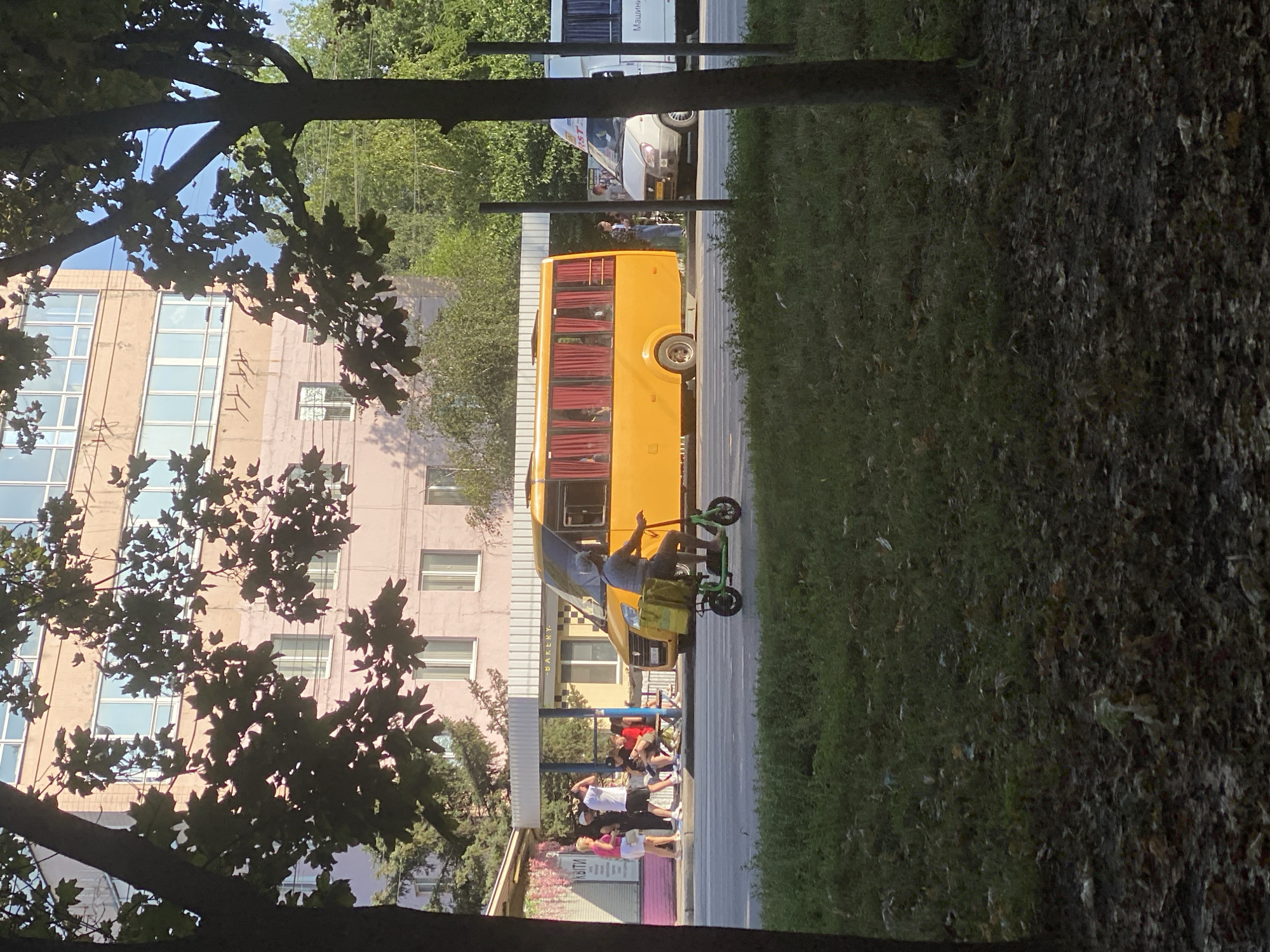 In Poltava, Ukraine, on a sunny day, a man rides a green electric scooter with a yellow insulated delivery bag on the front. He is wearing a gray t-shirt, shorts, and a cap, and is traveling on a paved road. Behind him, a bright yellow minibus is parked at a bus stop, its red curtains drawn. A white van is also visible nearby. Several people are standing and sitting at the bus stop. In the foreground, the road is bordered by a grassy area and trees. The background features a multi-story building with light-colored facade and windows.