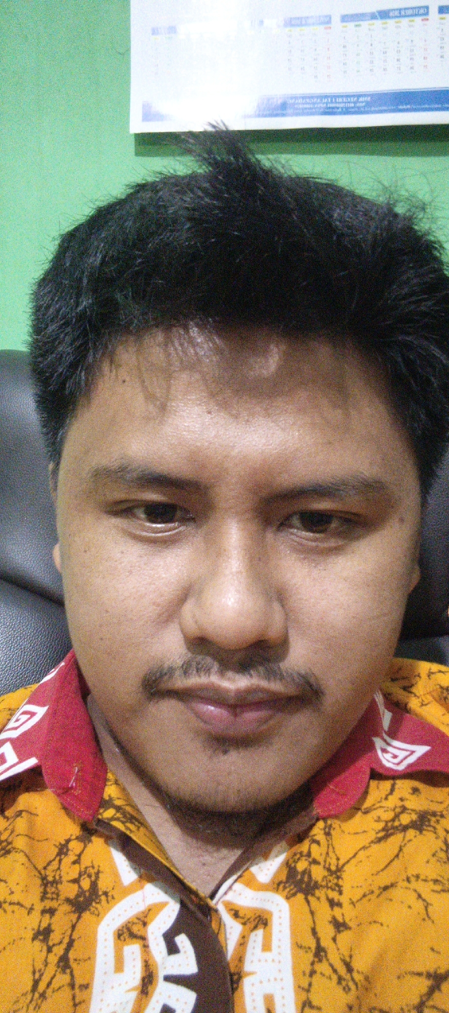 A medium shot selfie is taken of a man's face, from the chest up. The man has dark, messy hair, dark brown eyes, a mustache, and a small amount of stubble on his chin. He is wearing a yellow shirt with a brown, abstract pattern and a red collar with white trim. The background appears to be a light green wall with a calendar hanging on it. The image does not provide any specific clues about its location within Sukabumi, Indonesia.