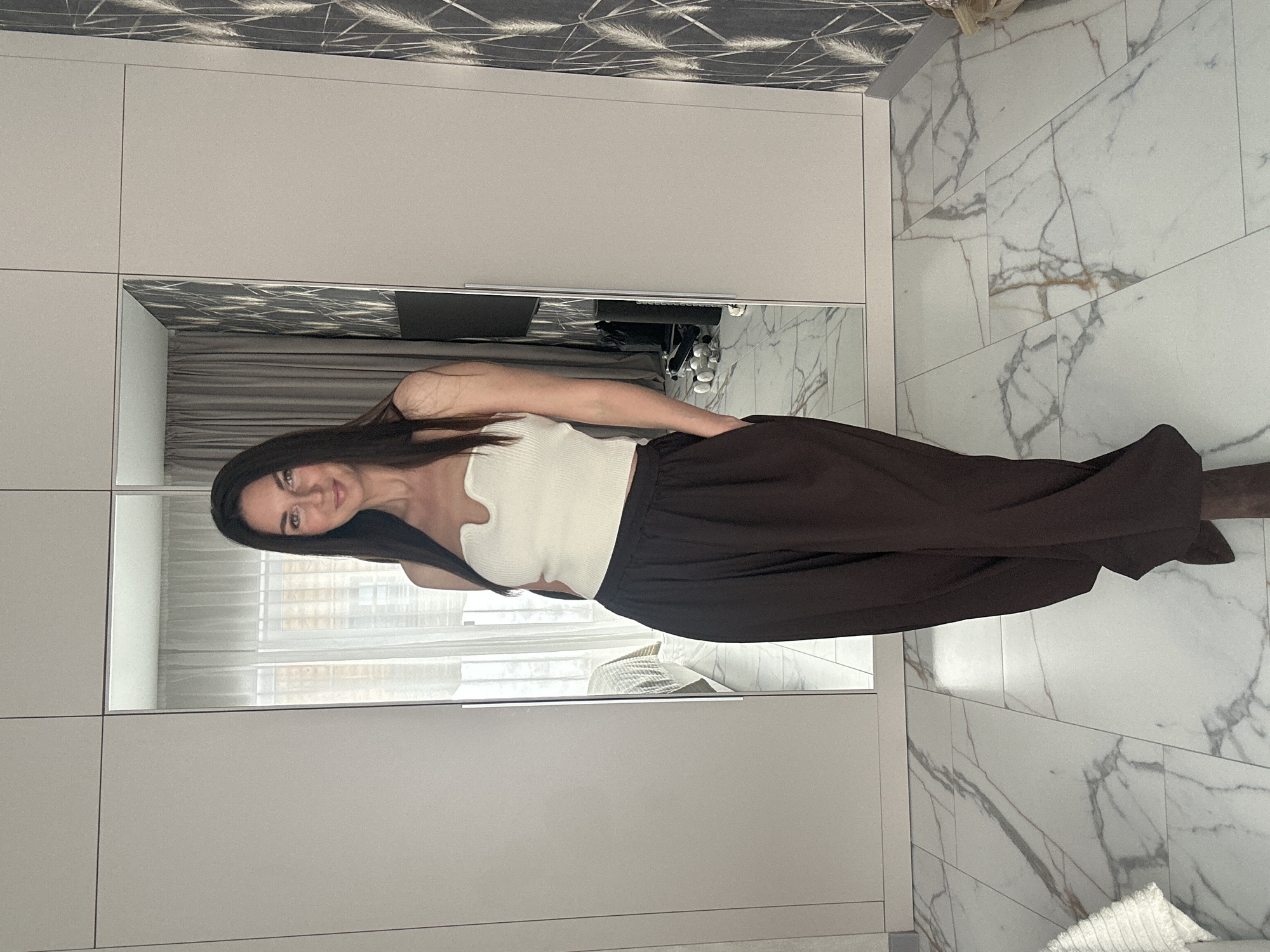 The scene depicts an adult female inside a modern room located in the city of Chaikovsky, Russia. The female, with long, dark brown hair and fair skin, stands centrally, facing the camera with a slight smile. She is dressed in a white, ribbed, strapless crop top with a V-neckline and dark brown, wide-leg trousers, with her right hand placed in a trouser pocket. Dark footwear is partially visible below the trousers. The floor is covered with white marble-patterned tiles featuring grey and brown veining; a small section of a light-colored, textured rug is visible in the bottom-left corner. Behind the individual is a light grey modular wardrobe unit. Two central doors of this unit are mirrored, reflecting parts of the room. The left mirror shows a bed with a light-colored duvet and a window with sheer white curtains. The right mirror reflects a dark grey curtain or wall, a wall-mounted television screen, and a black bag with several white, rounded objects on the floor beneath the television. To the right of the wardrobe, a wall is adorned with dark grey wallpaper featuring a pattern of light, elongated, feather-like designs.