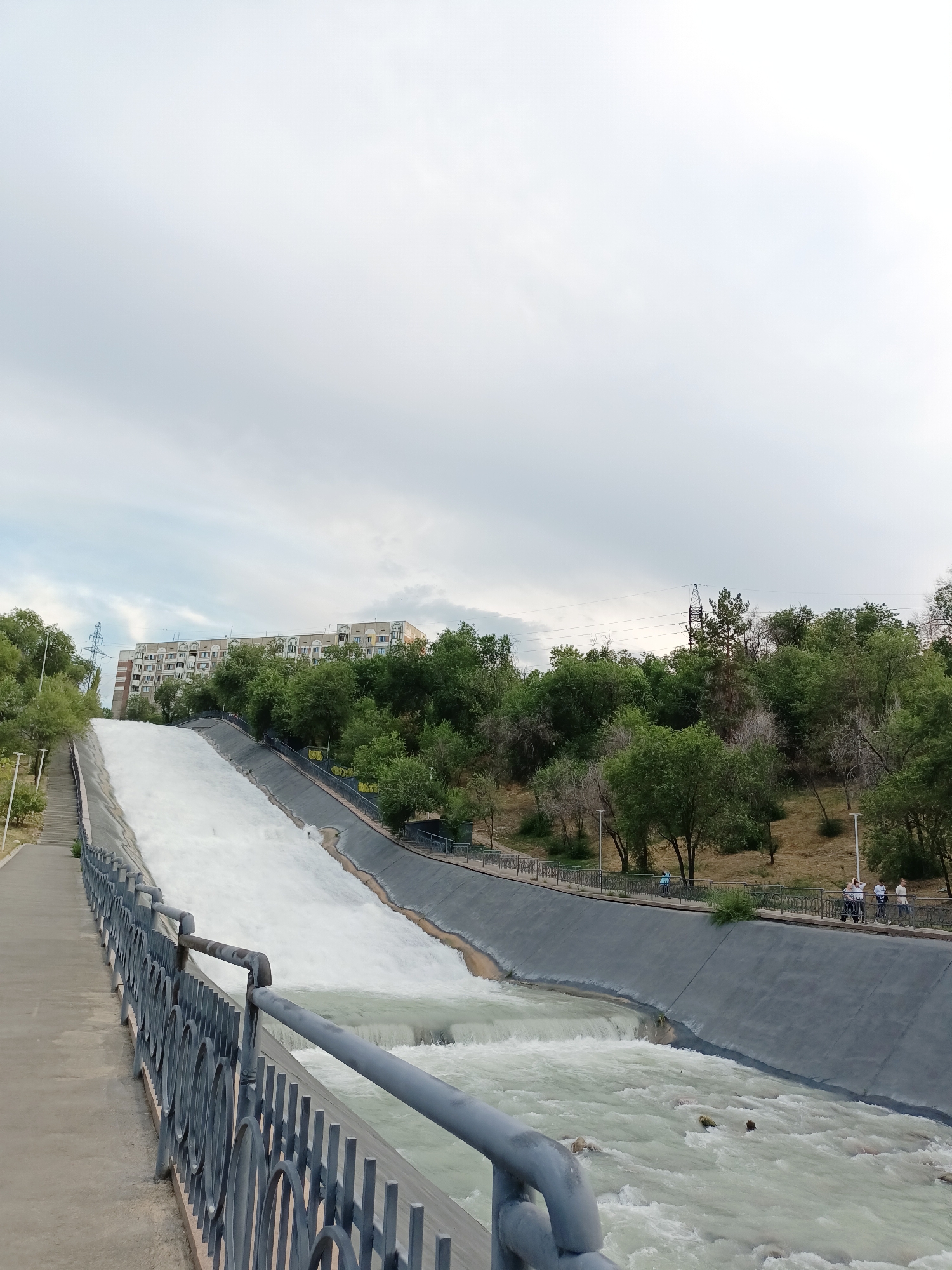 This image captures a view of the large concrete spillway of the Almaty Dam, located in the city of Almaty, Kazakhstan. The spillway is actively channeling a significant amount of white water down its steep incline, creating a dynamic and powerful visual. On the left side of the spillway, a paved walkway with steps leads upwards, bordered by a decorative metal railing. The right side of the spillway is lined with a concrete embankment, also featuring a railing and walkway.
A line of green trees and foliage borders the area, with more dense vegetation on a hillside in the background. Behind the trees, an apartment building with multiple floors and balconies is visible. Power lines and a tall electrical pylon are also present in the distance. Several people are seen walking along the embankment walkway, observing the water. The sky is overcast with light clouds.