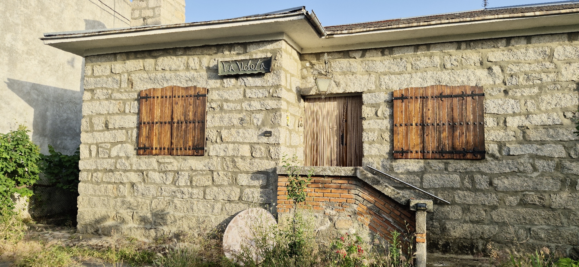 A quaint, single-story stone building with a sloped roof is viewed at eye-level in a rural setting. The facade is constructed from rough-hewn granite blocks of varying sizes, with visible mortar. To the left, a wooden shutter with vertical planks and black metal accents covers a window. Above the shutter, a dark metal sign with white, calligraphic lettering reads "LA VELATA." To the right of the sign, a door with a beaded curtain hangs as a screen. To the right of the door, another identical wooden shutter covers a window. A small brick staircase with a stone railing leads up to the doorway. Overgrown vegetation, including a bush with white flowers and tall grasses, surrounds the base of the building and extends to the left where a chain-link fence is partially visible. The background shows a portion of an adjacent building with a lighter, concrete-like finish. The sky above is a clear, pale blue. The scene is illuminated by natural sunlight, casting shadows on the facade and the adjacent building.