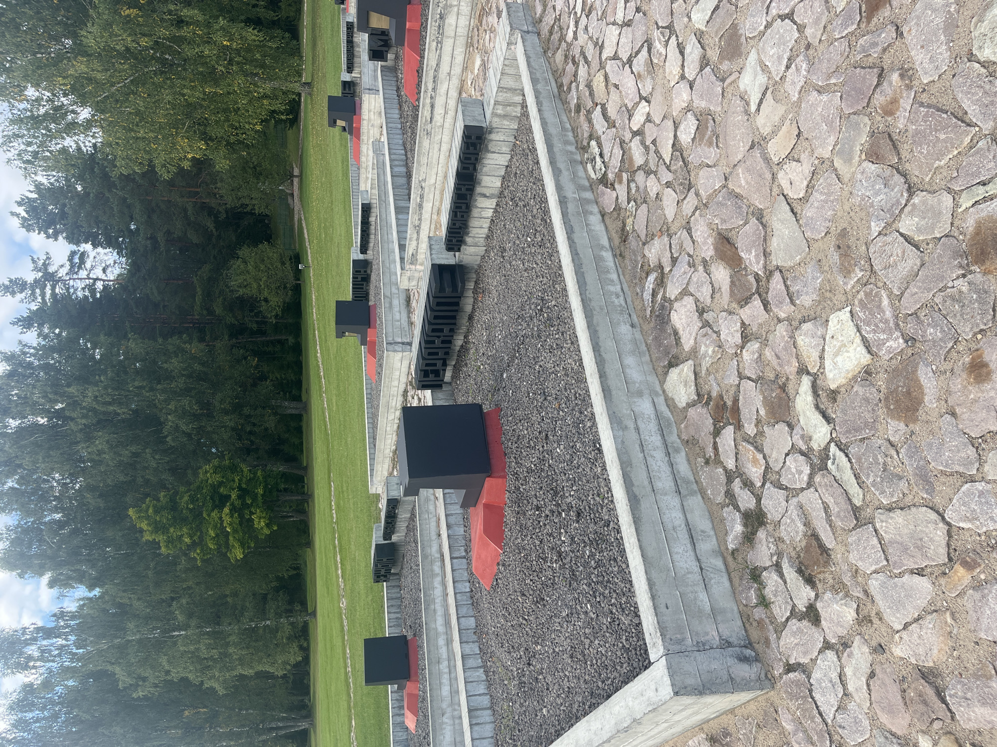 The image depicts a memorial site with several raised concrete beds filled with dark gravel. Each bed contains a black, cubical monument with a red base. Some of the monuments have black lettering on their sides. The memorial is set in a landscape with a mown lawn and a backdrop of tall, mature trees, including birches and pines. The ground in the foreground is paved with irregular, flat stones. The lighting suggests it is daytime with some cloud cover. The style of the memorial, with its stark cubical forms and engraved lettering, suggests a historical or commemorative purpose. The location is identified as Каменскі сельскі Савет, Belarus.