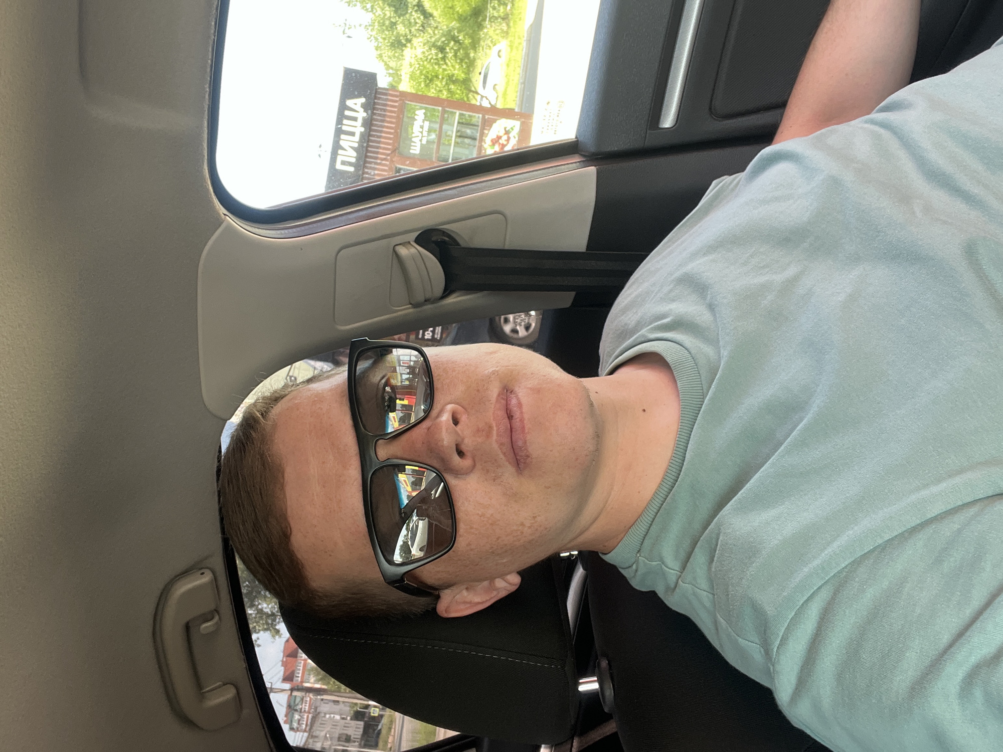 A man wearing a light teal t-shirt and dark sunglasses is seated in the passenger seat of a car. The interior of the car is visible, including the headrest and a seatbelt. Through the passenger side window, a building with signage that reads "ПИЦЦА" (Pizza) and "ШАУРМА" (Shawarma) is visible. The background shows greenery and a white car. The man's face is partially obscured by his sunglasses, and he appears to be looking forward. The image was taken during the daytime, under clear skies.